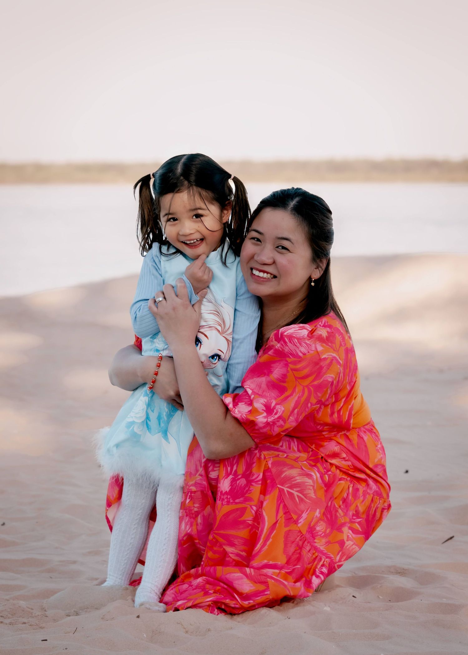 A mother and child in bright pink and blue outfits share a joyful moment playing together on a sandy beach.
