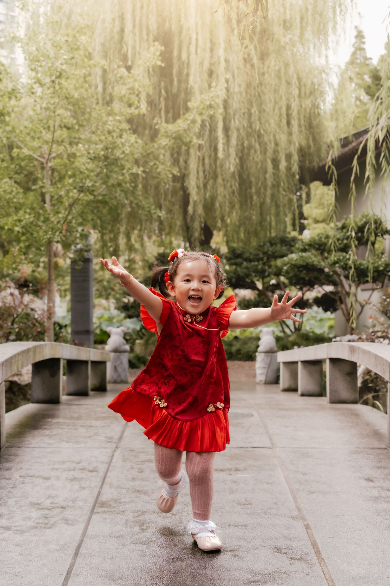 A joyful toddler girl in a red Chinese dress runs with arms outstretched in a lush garden setting.