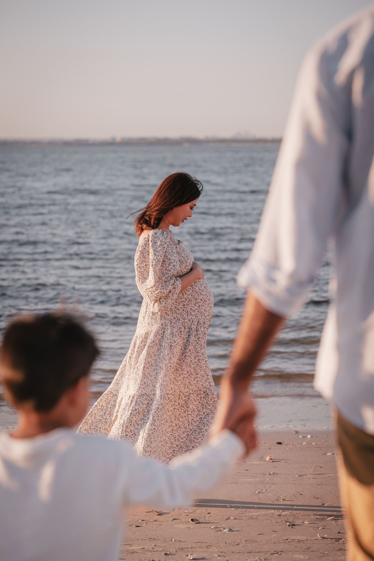 A person in a white floral dress walks along a sandy beach at sunset with the ocean in the background.