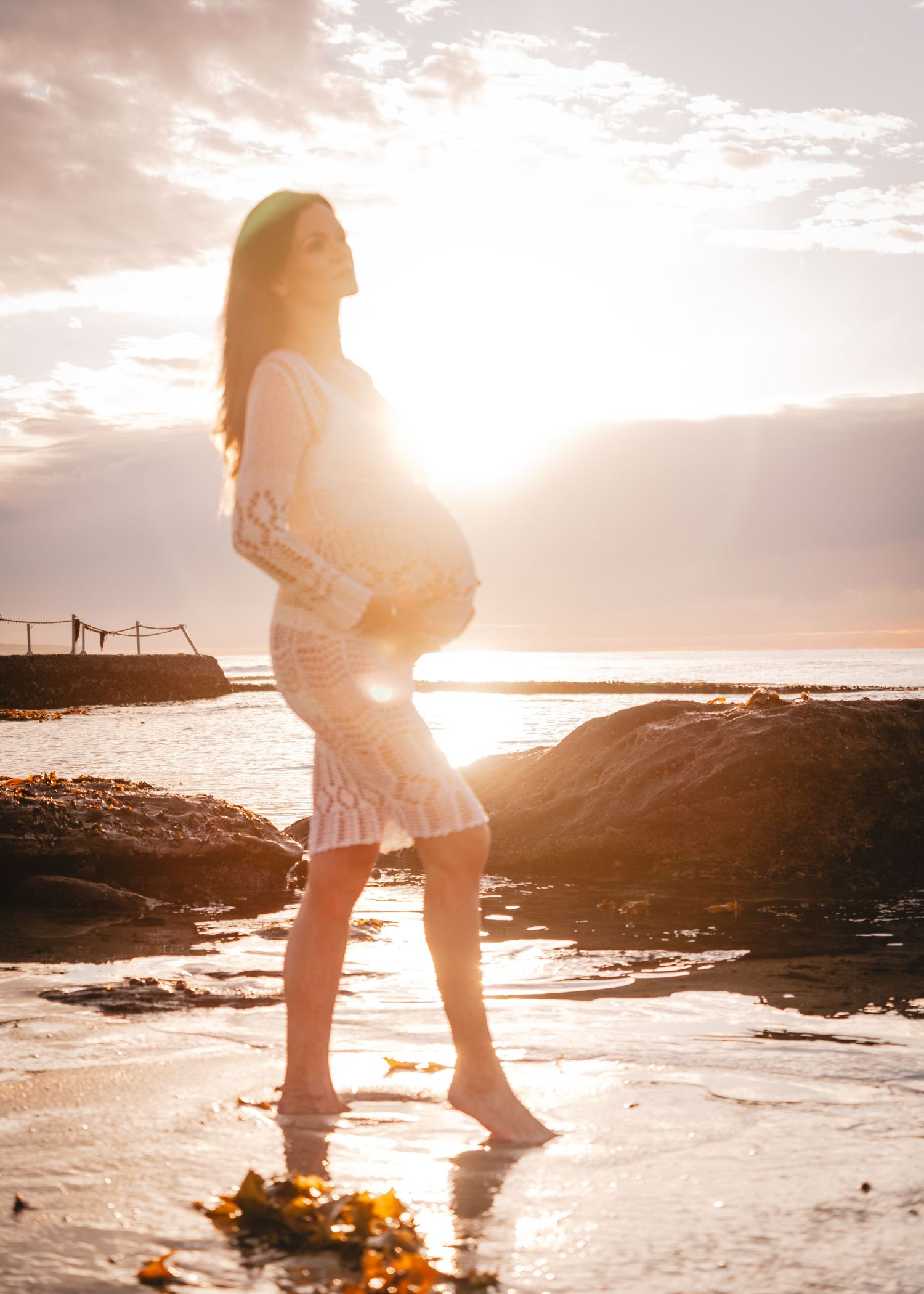 Silhouetted pregnant woman stands on beach at sunset with sun rays creating golden glow on wet sand.