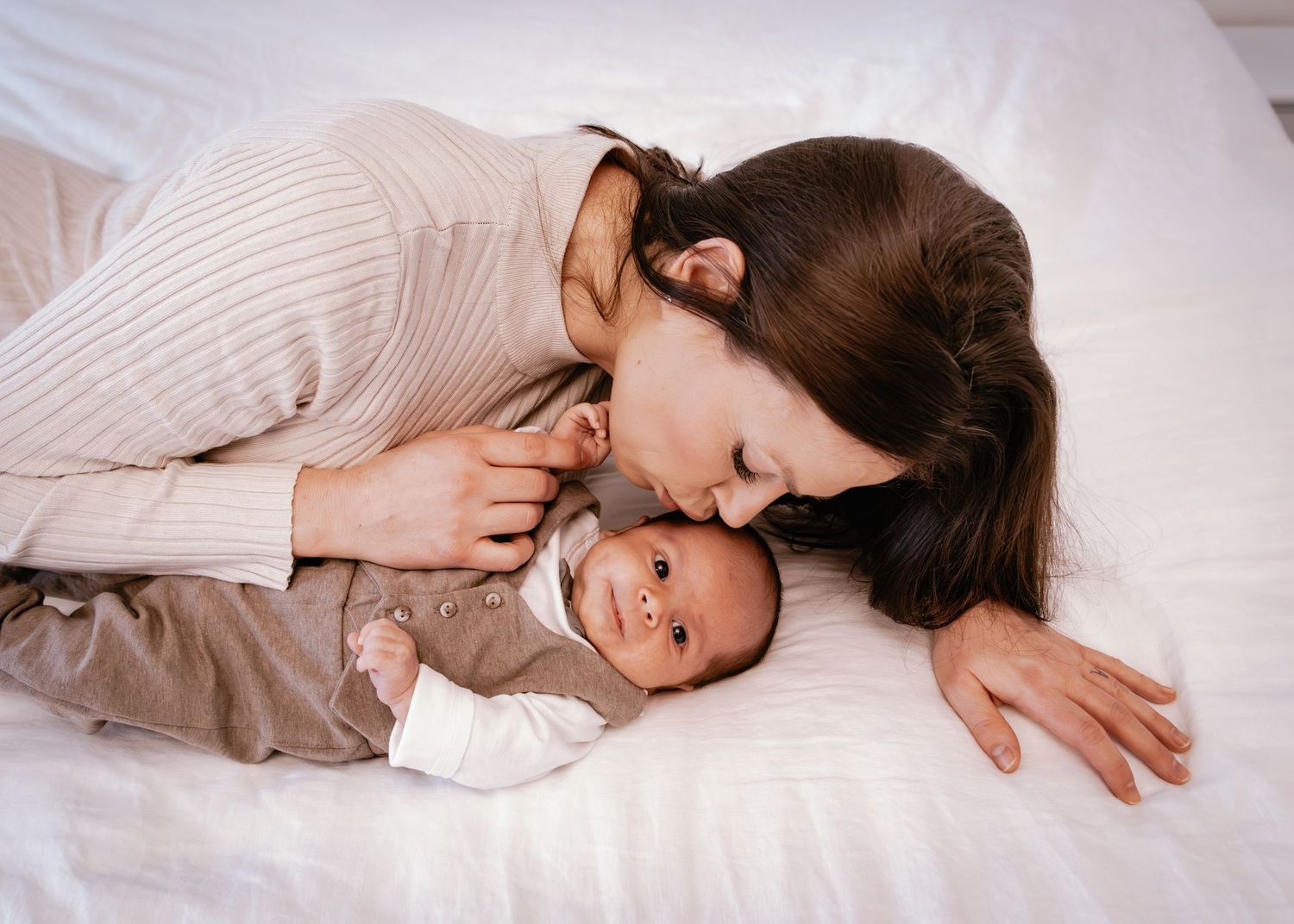 A tender moment captured as a parent in a cream sweater cuddles with their smiling baby on white bedding.