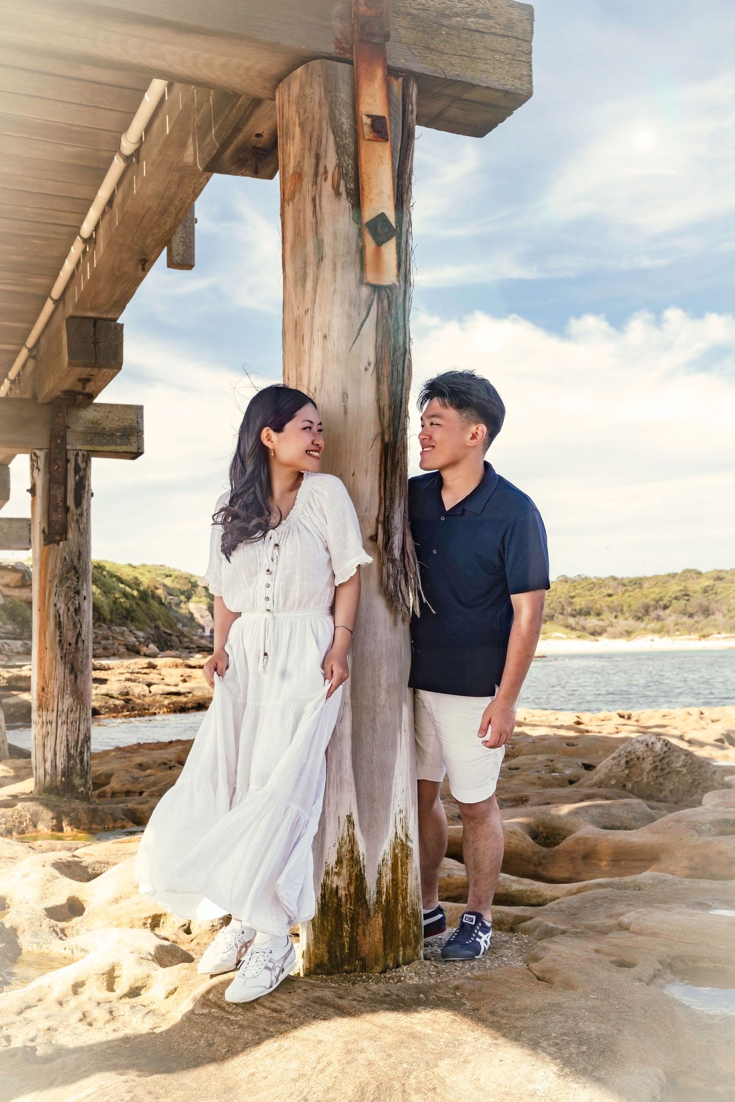 A couple shares tender moments under a wooden pier on a sandy beach during sunset.