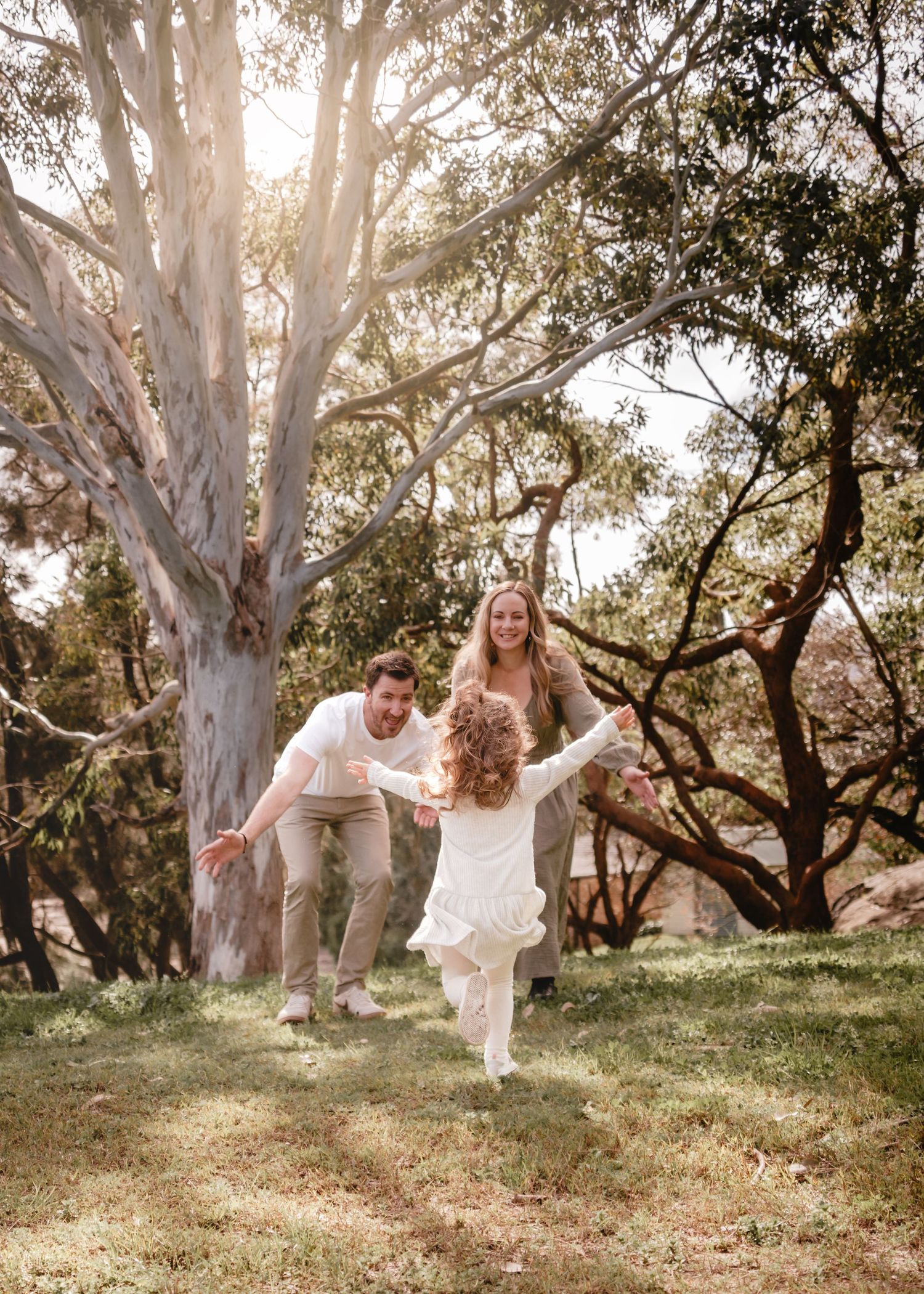 Family playing outdoors near large eucalyptus tree, parents chasing laughing child in white dress on green grass.