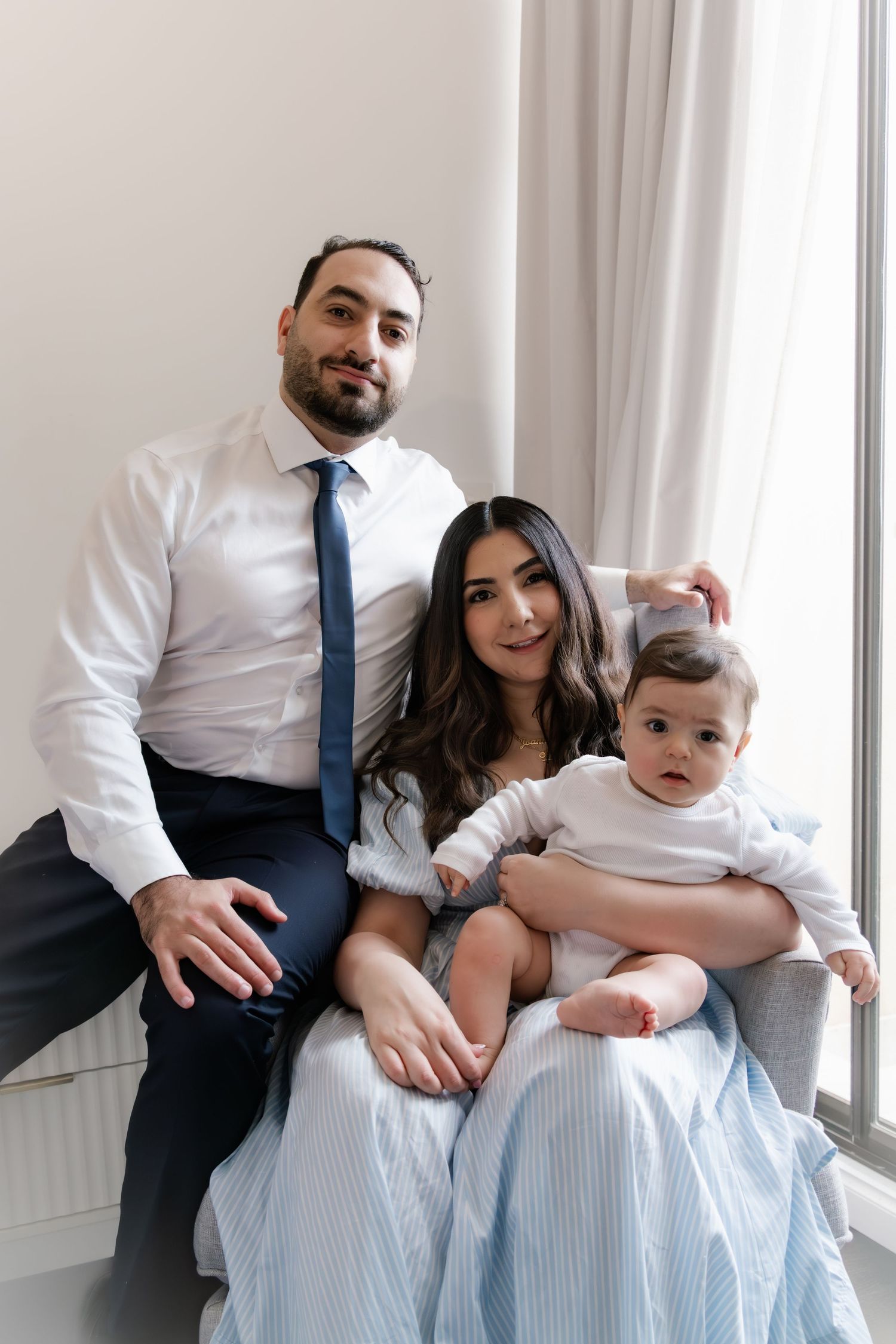 A family portrait of a man in a white shirt and blue tie, a woman in a blue dress, and their baby in white.