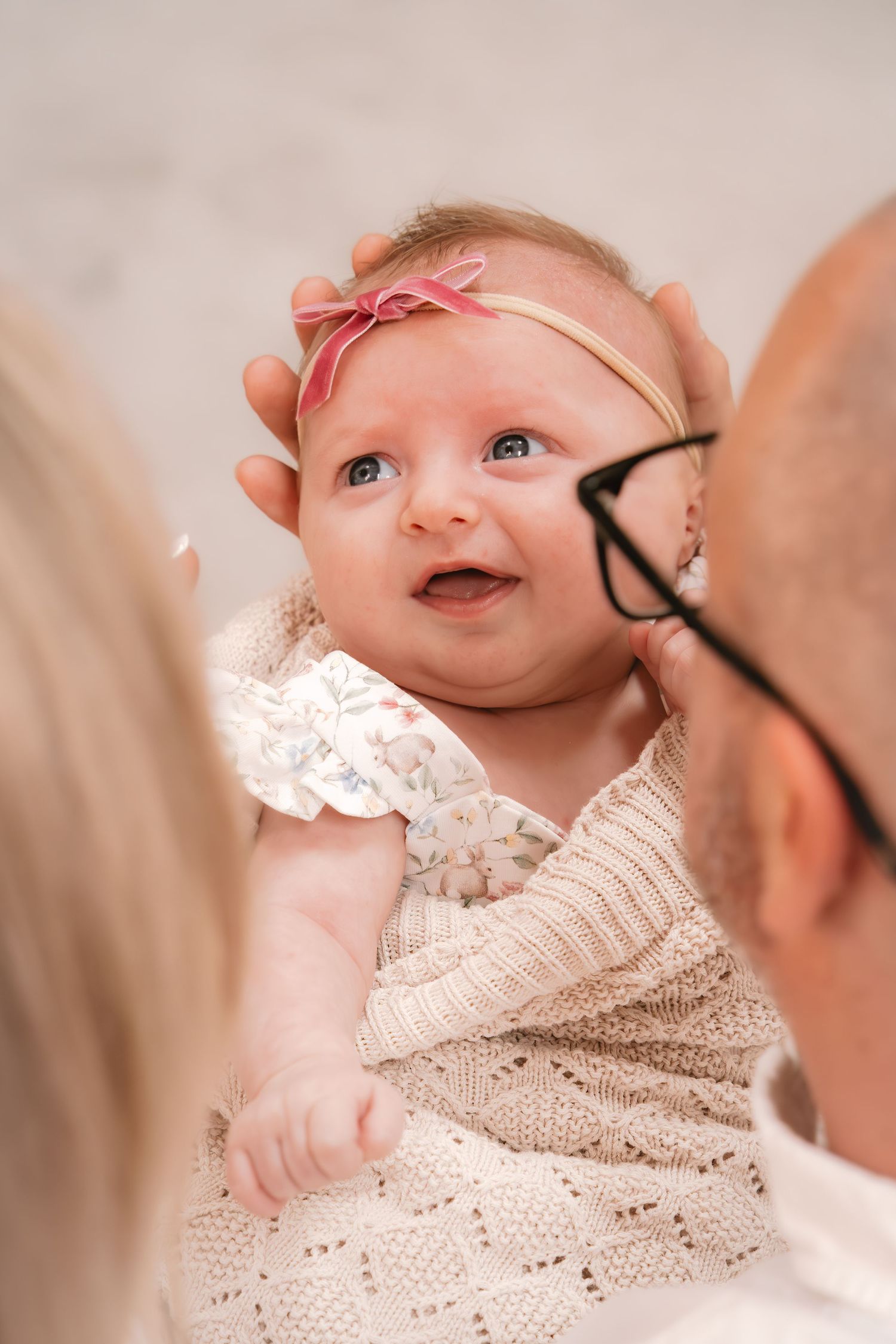 A baby wearing a pink headband and white lace dress smiles while being held.