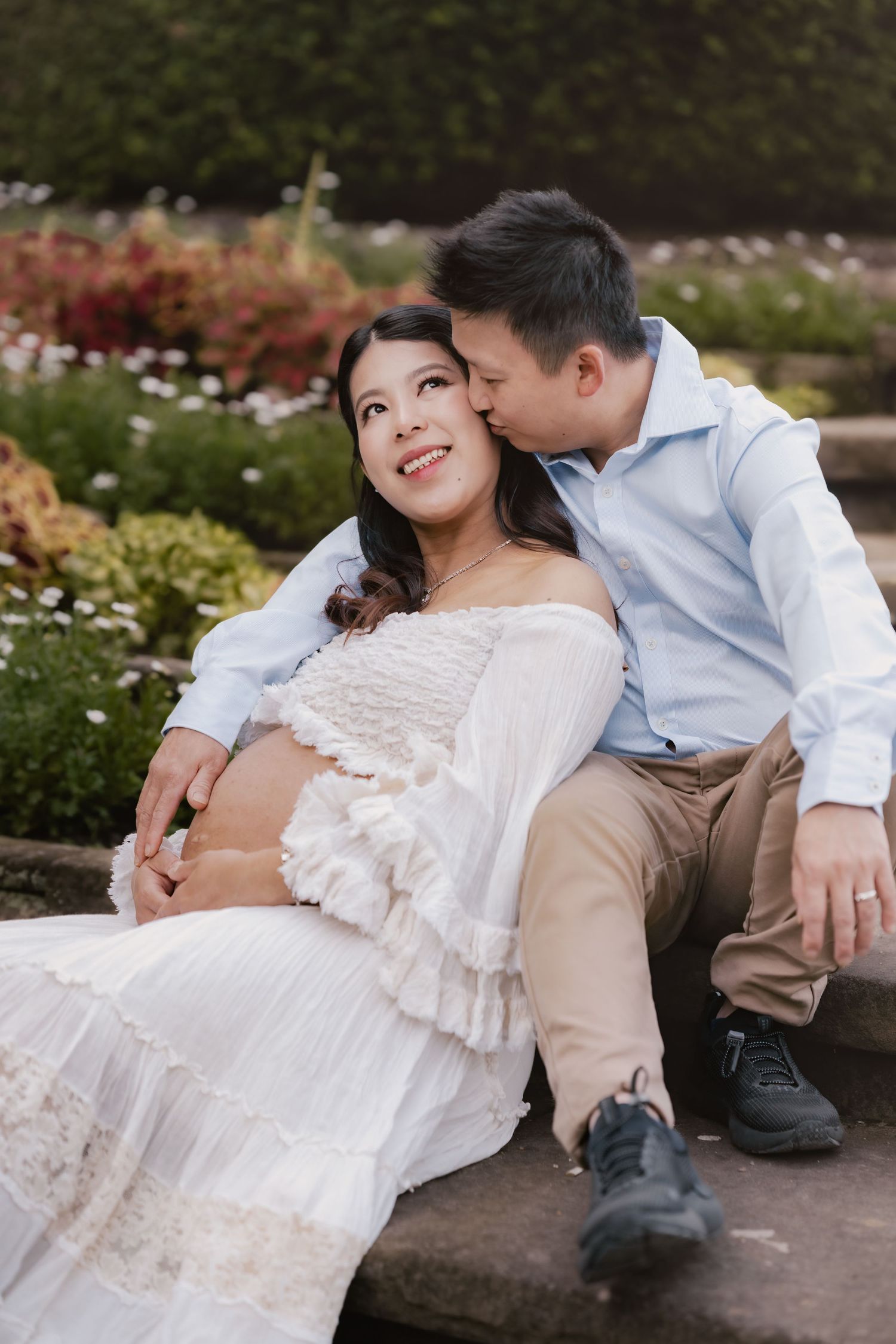 A man kisses a smiling pregnant woman in a white dress as they sit together in a blooming garden.