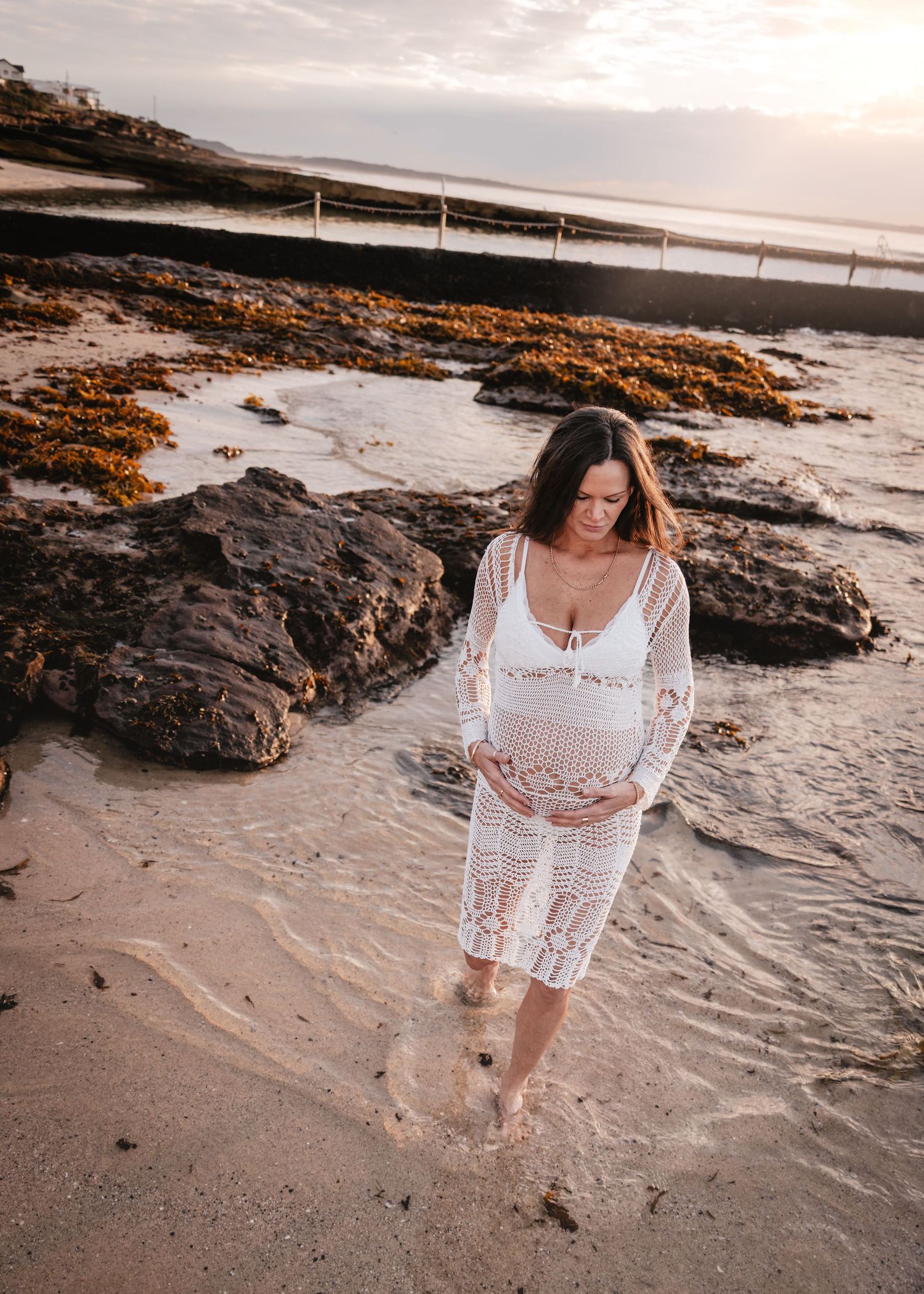 A person in a white crochet cover-up walks along a rocky beach at sunset creating a dreamy coastal scene.