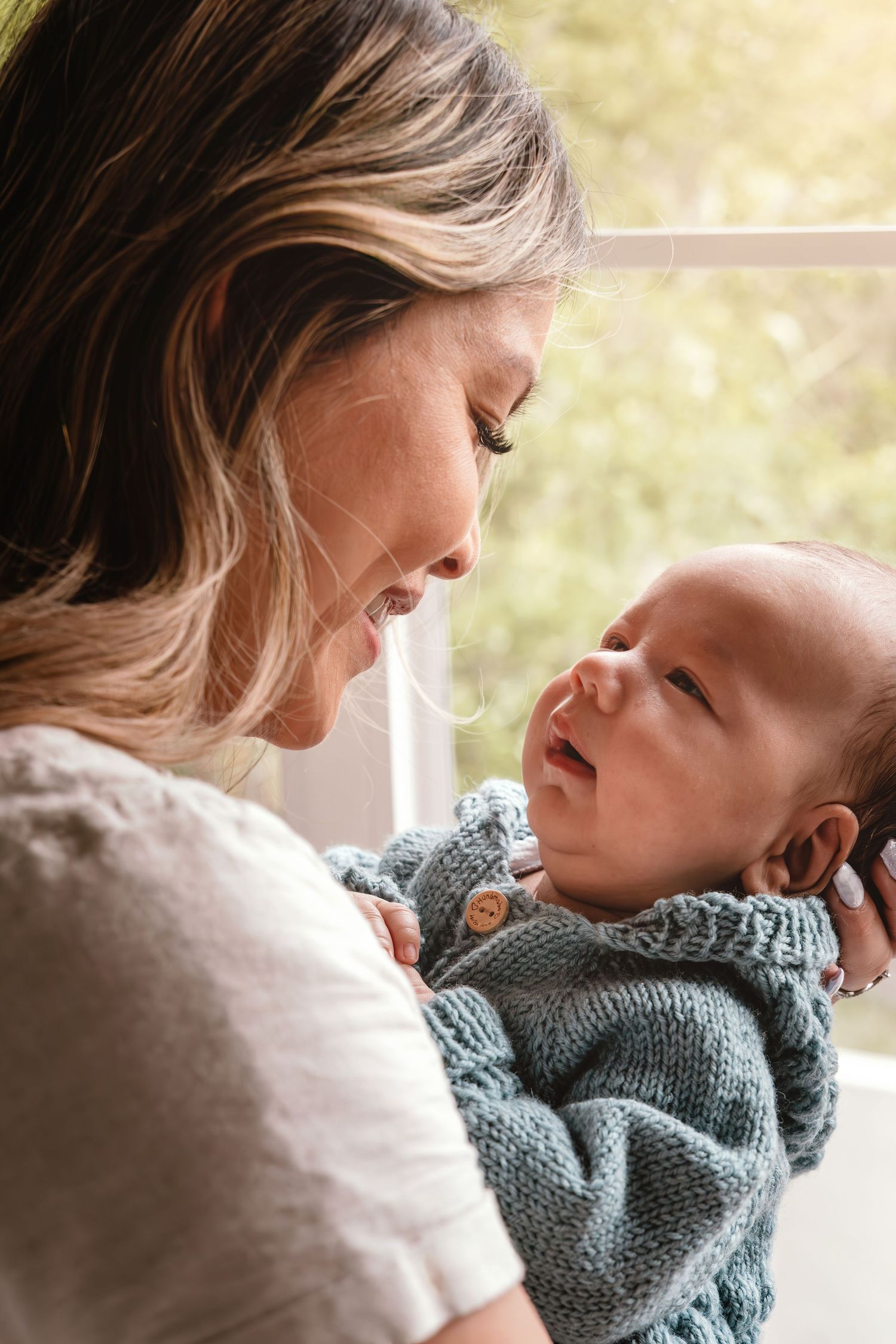 A smiling mother tenderly holds her newborn baby in a knit outfit near a sunlit window.