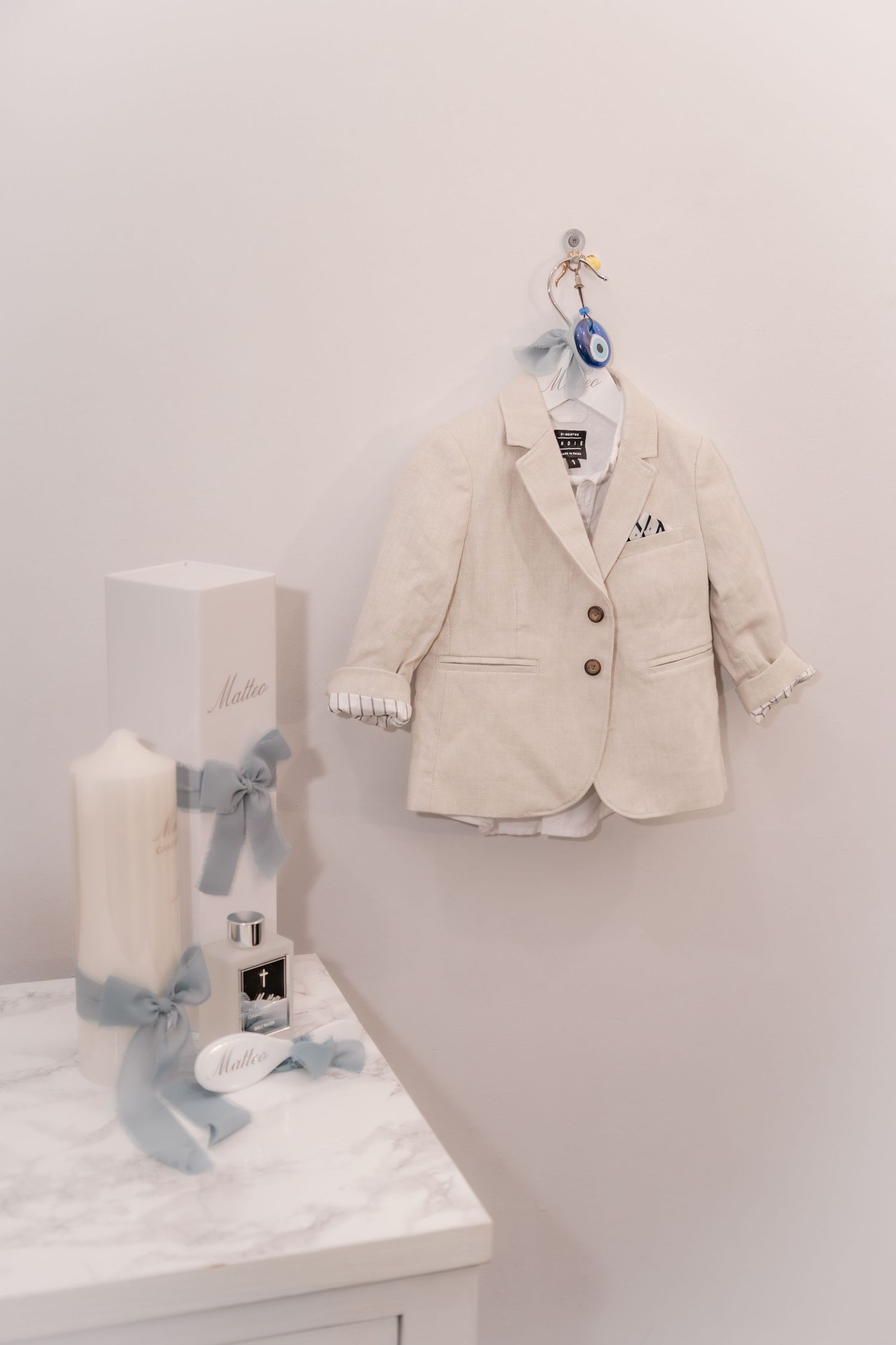 Small beige baby boy baptism suit on hanger beside white candles with blue ribbons on marble dresser.