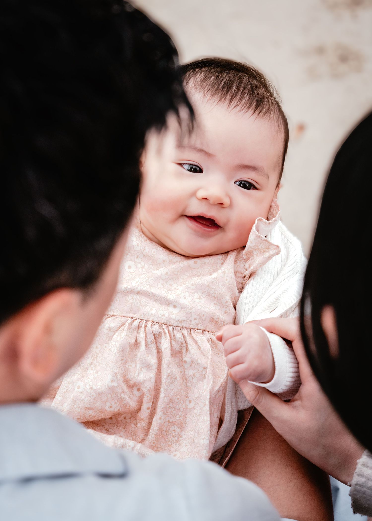 A smiling baby wearing a pink polka dot dress looks up while being held by someone in a blue shirt.
