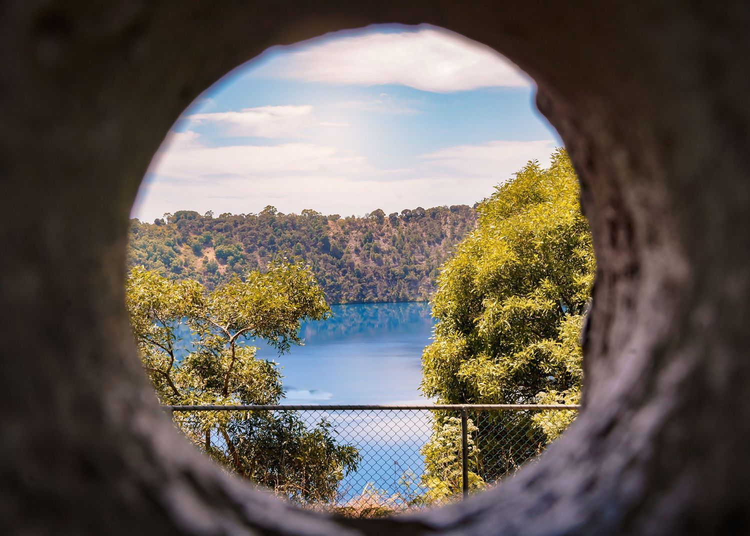 Deep blue lake waters and tree-covered hillsides seen through a dark tunnel opening against a bright sky.