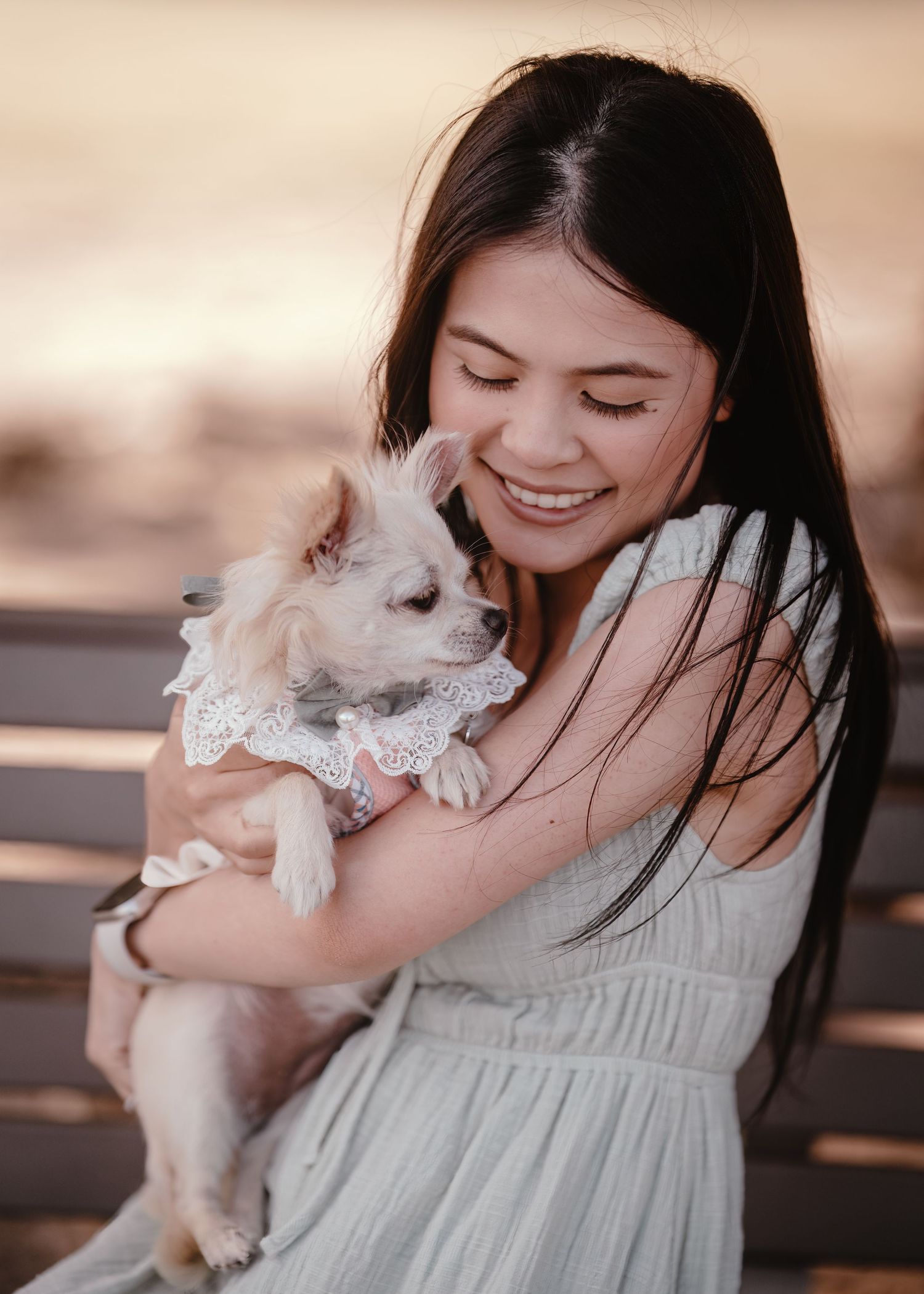 A person in a light dress smiles while holding a small white dog wearing a lace collar in a warm outdoor setting.