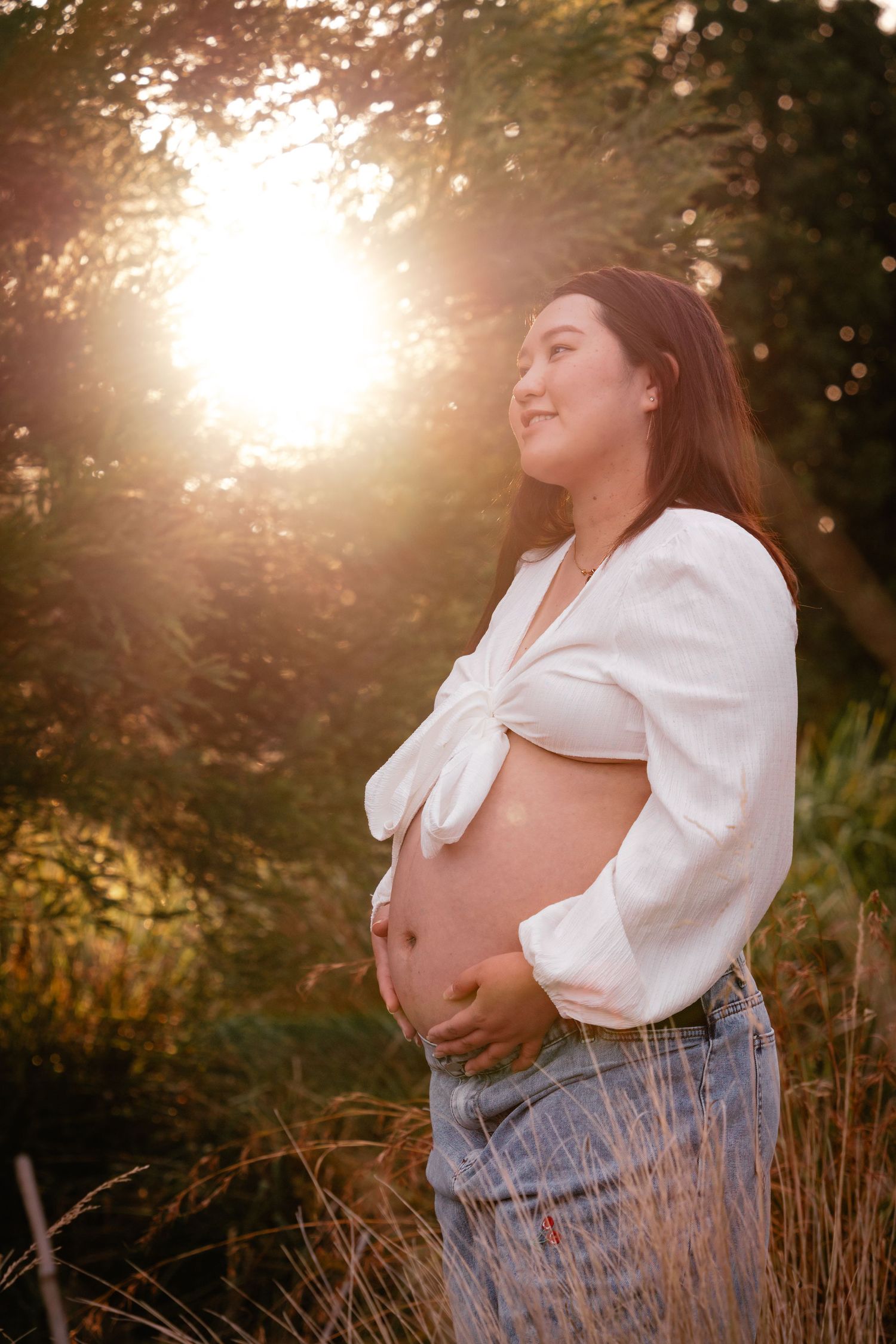 A maternity photo series showing a pregnant person in a white top and jeans standing in golden sunlit field.