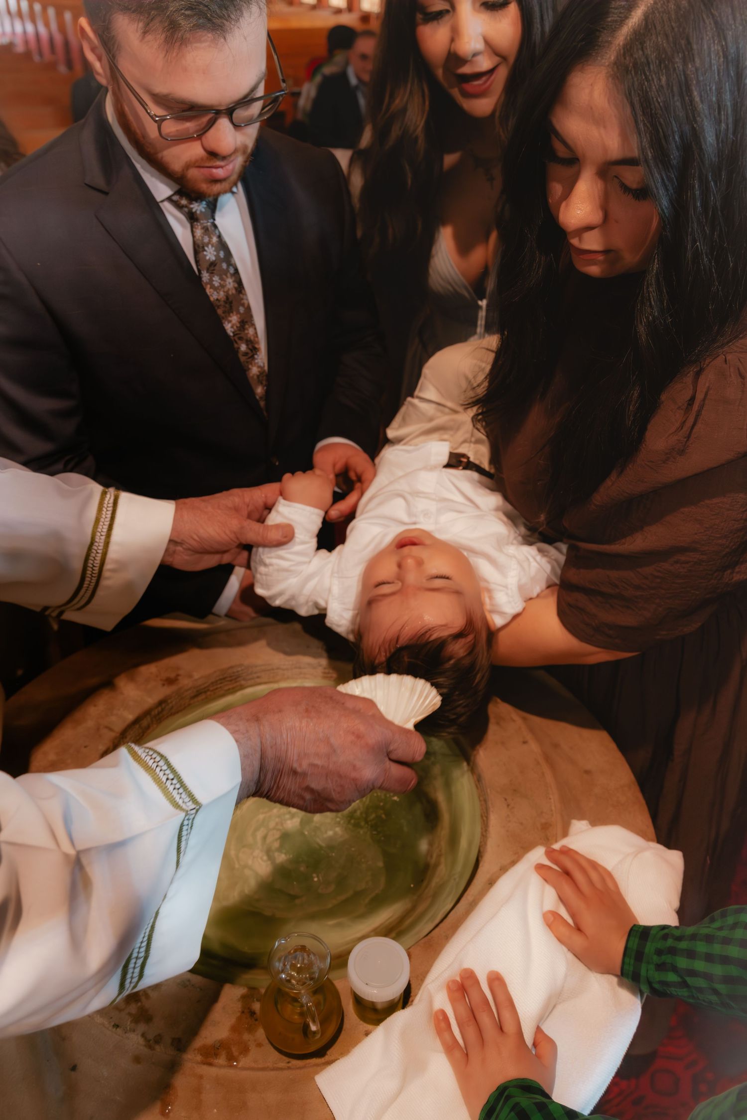 A Catholic priest baptizes an infant at a stone font, surrounded by proud parents in formal attire.