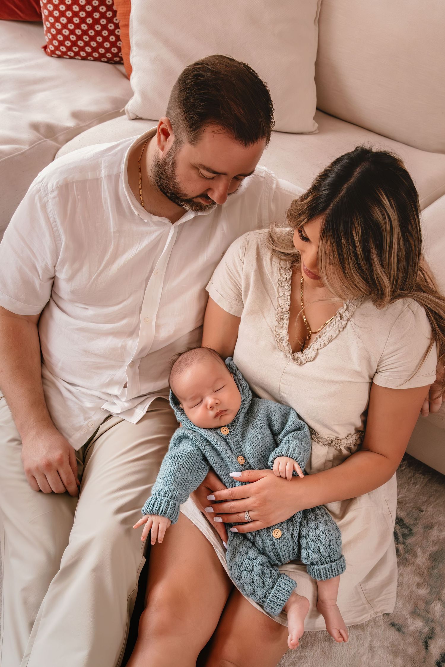 A peaceful family moment as parents in white shirts cuddle with their sleeping baby wearing a blue knit outfit on the couch.