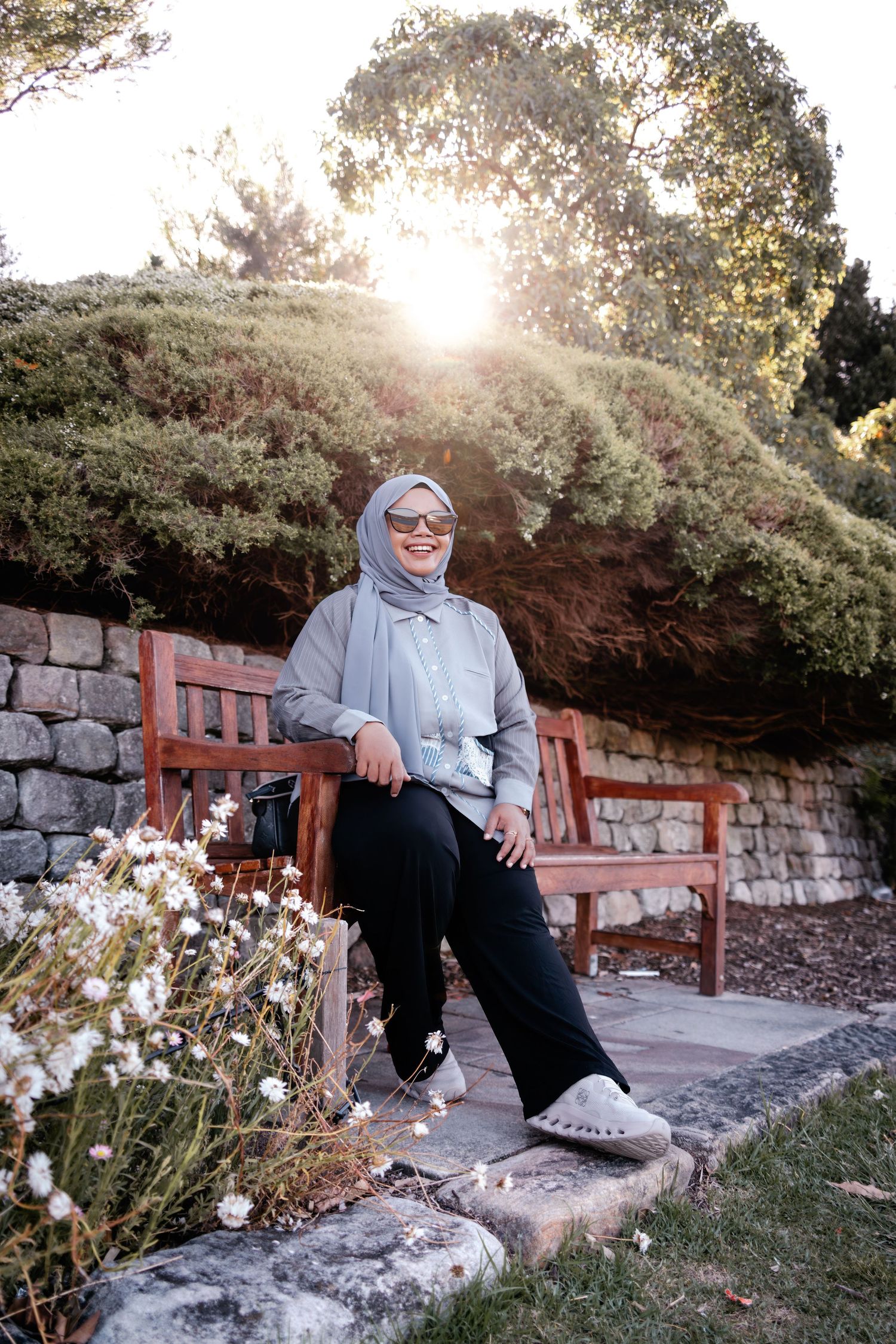 A person in a gray hijab and casual attire sits on a wooden bench in a garden during golden hour sunset.