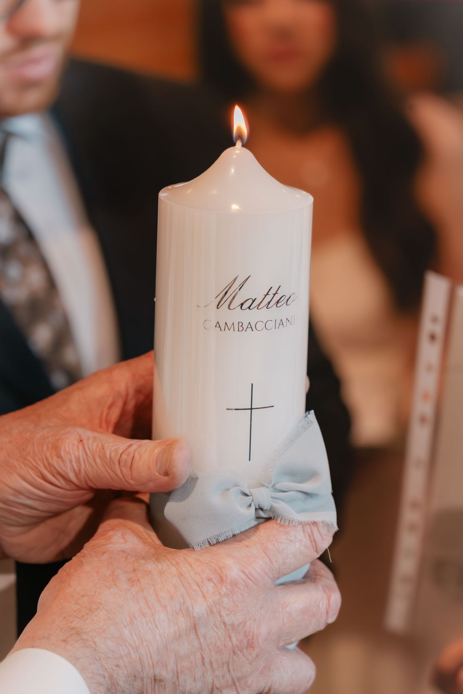 Hands holding a lit white baptism candle with cross and name Matteo Gambacciani, decorated with a blue bow.
