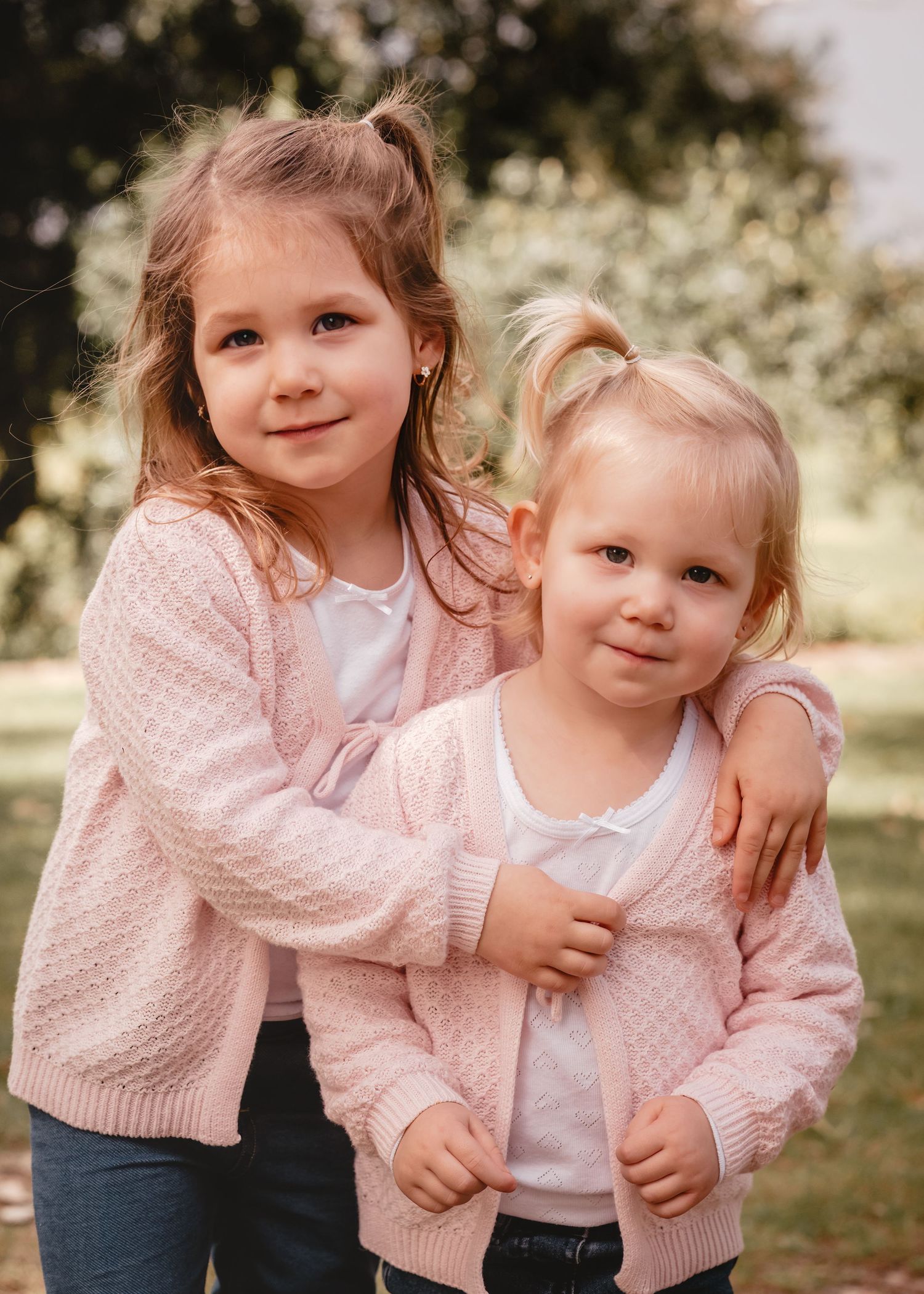 Two children wearing matching pink knit cardigans and white tops pose together outdoors in a garden setting.