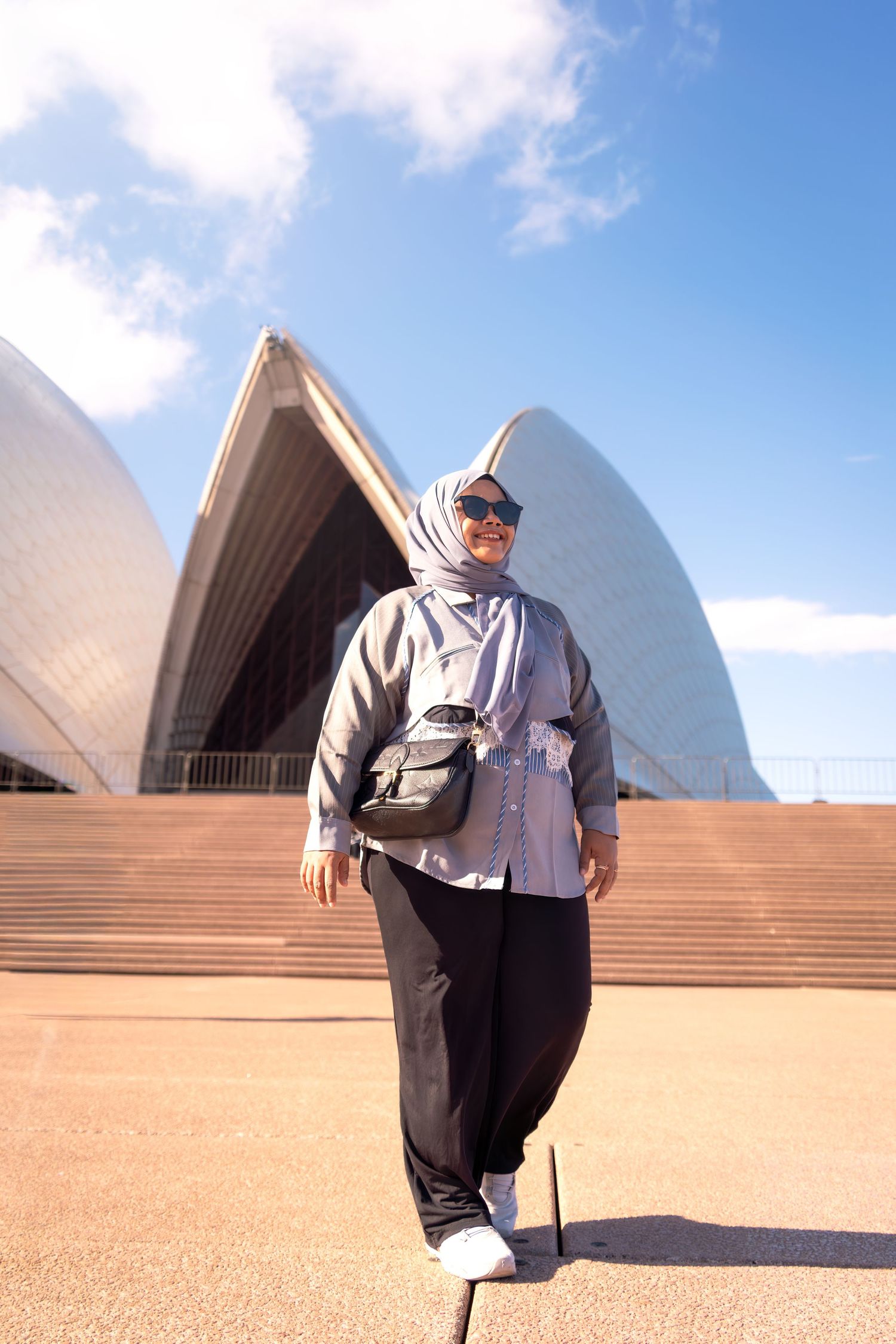 Tourist in casual attire poses on the steps of the iconic Sydney Opera House on a sunny day.