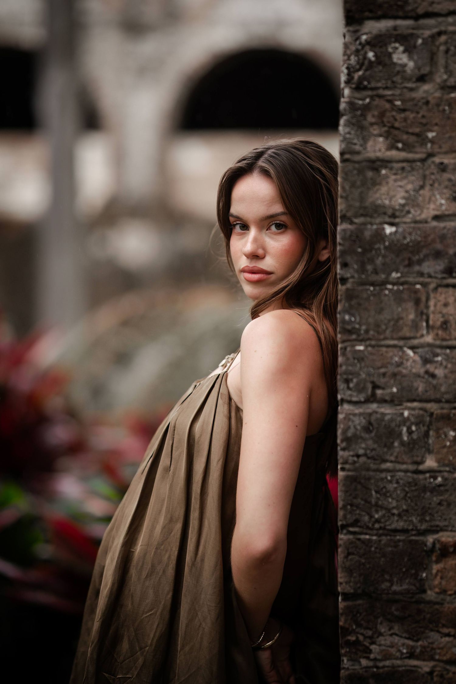 A woman in a brown sleeveless dress poses against a brick wall in a moody outdoor portrait series.