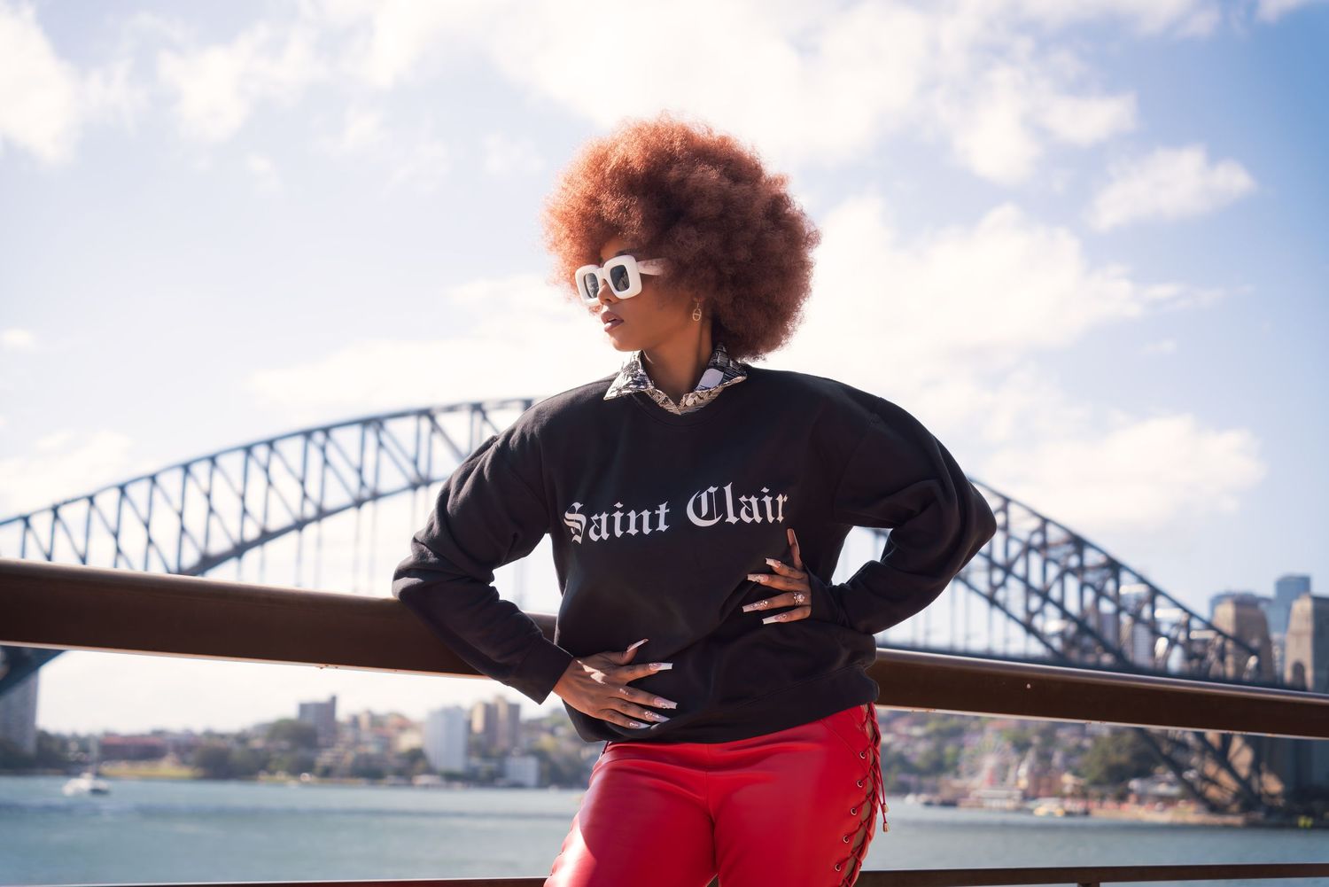 A person in a red dress poses in front of Sydney Harbour Bridge on a sunny day.