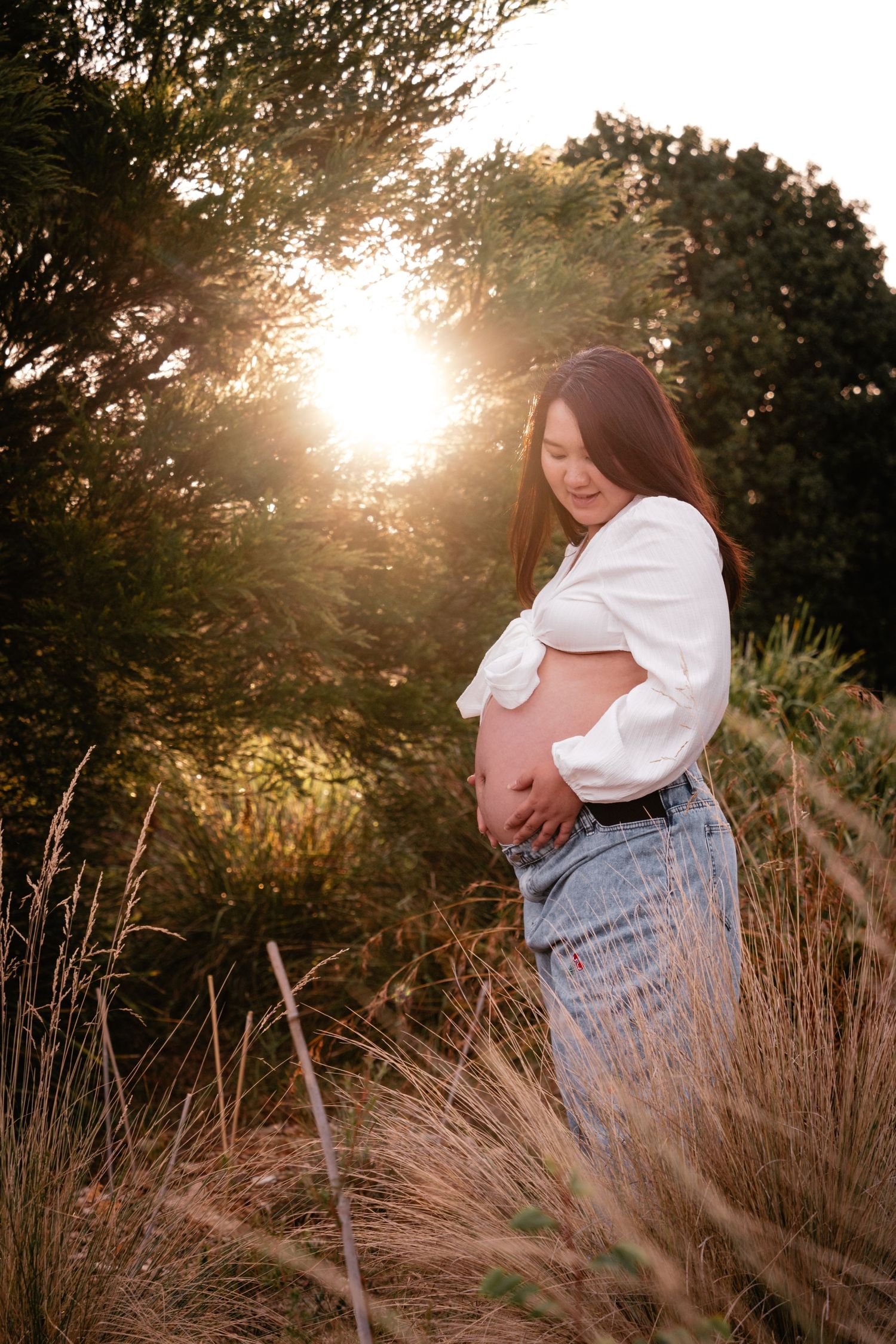 A maternity photo in golden sunlight shows an expectant mother in jeans and white top standing among tall grasses at sunset.