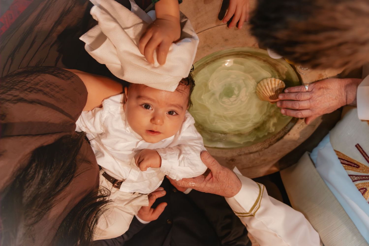 A baby in white clothing during a traditional baptism ceremony with adults holding a green ceremonial bowl overhead.