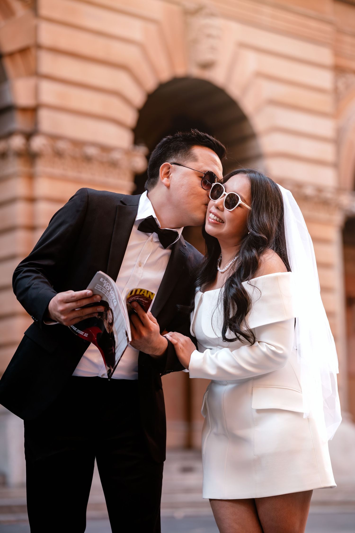 Asian couple in wedding attire, groom in black tuxedo kissing bride in white mini dress and veil, holding wine outside.