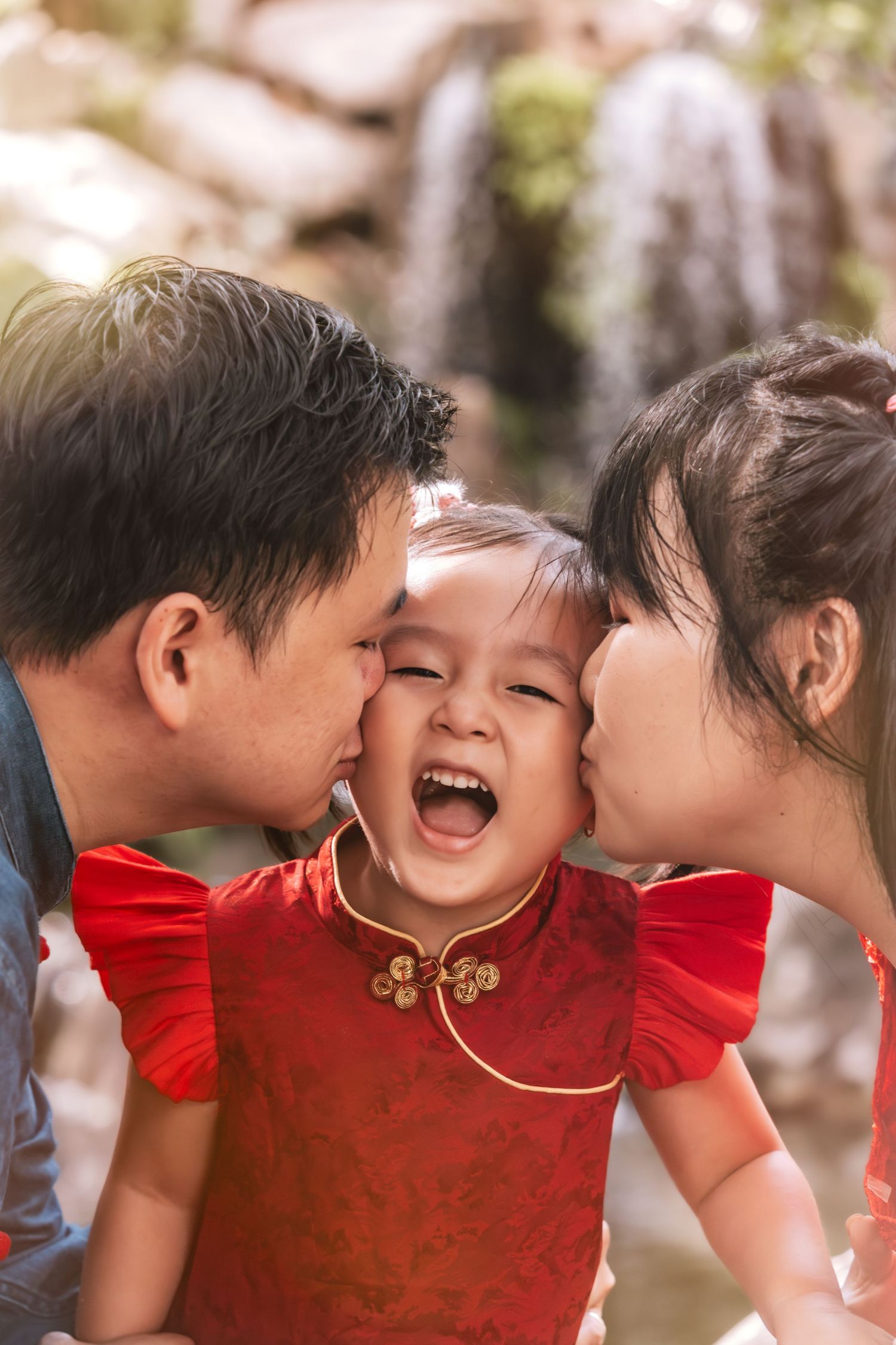 Asian parents kissing their laughing toddler girl in red cheongsam dress outdoors near a waterfall.