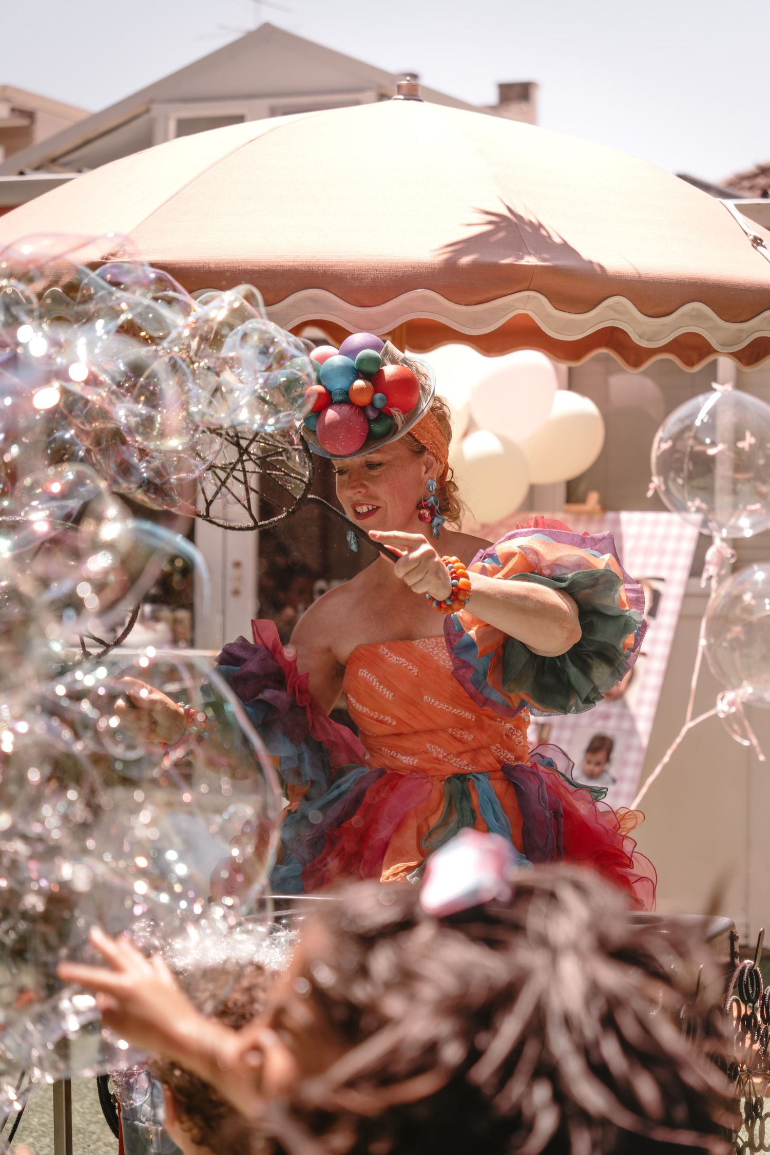 Colorful performer in festive costume blowing giant soap bubbles at an outdoor carnival event on a sunny day.