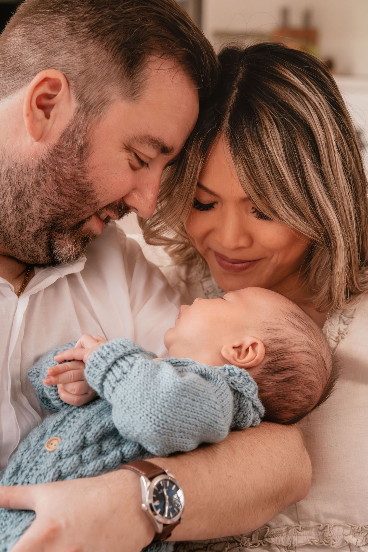 A tender family moment as parents lovingly gaze at their newborn baby wearing a soft blue knit sweater.