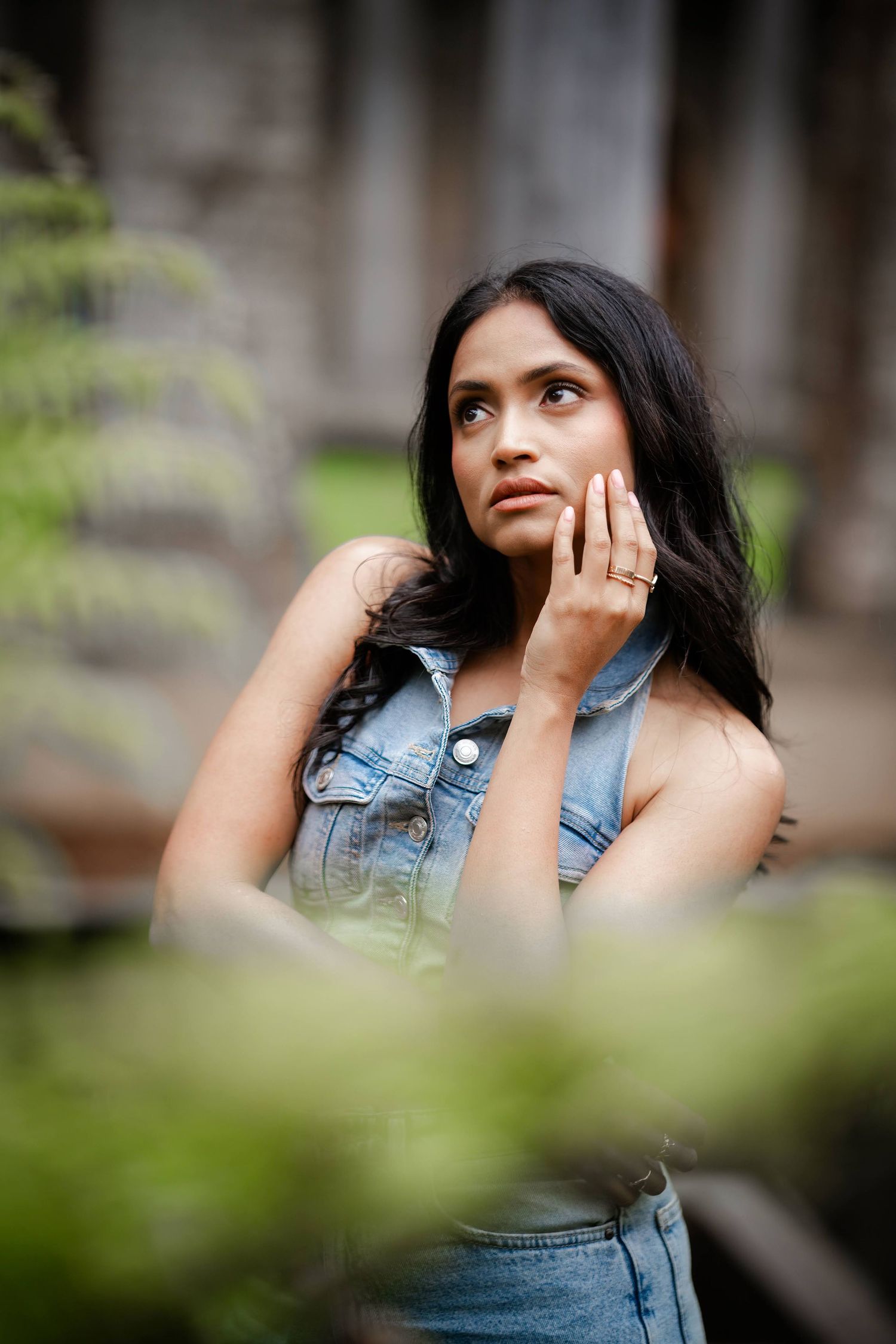 Woman in denim vest poses thoughtfully outdoors with greenery in foreground and soft bokeh background.