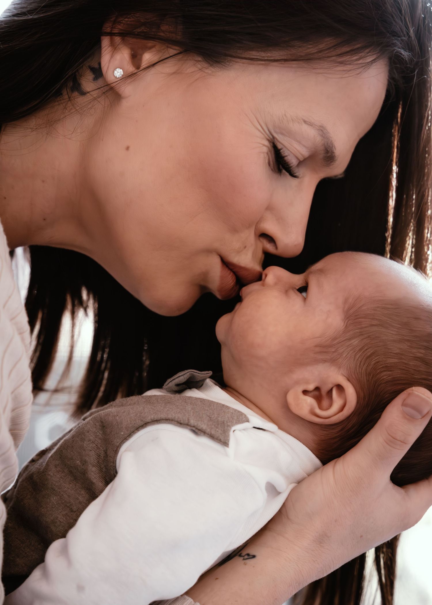 A tender moment captured between a mother and newborn baby as they share a gentle kiss in warm lighting.