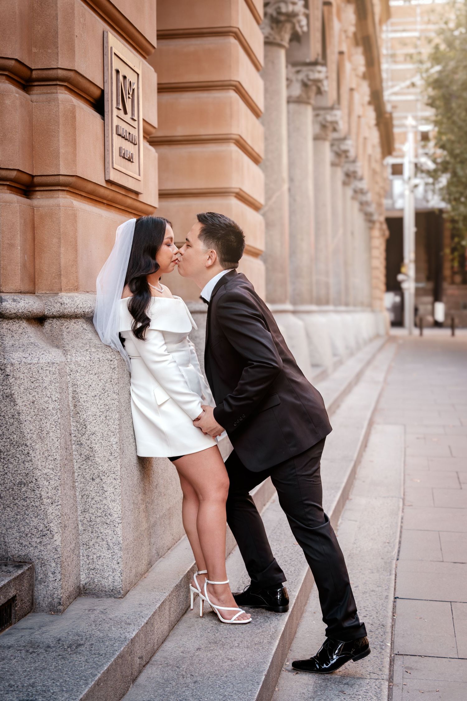 Bride and groom kissing against a historic stone building, bride in white mini dress with veil, groom in black suit.