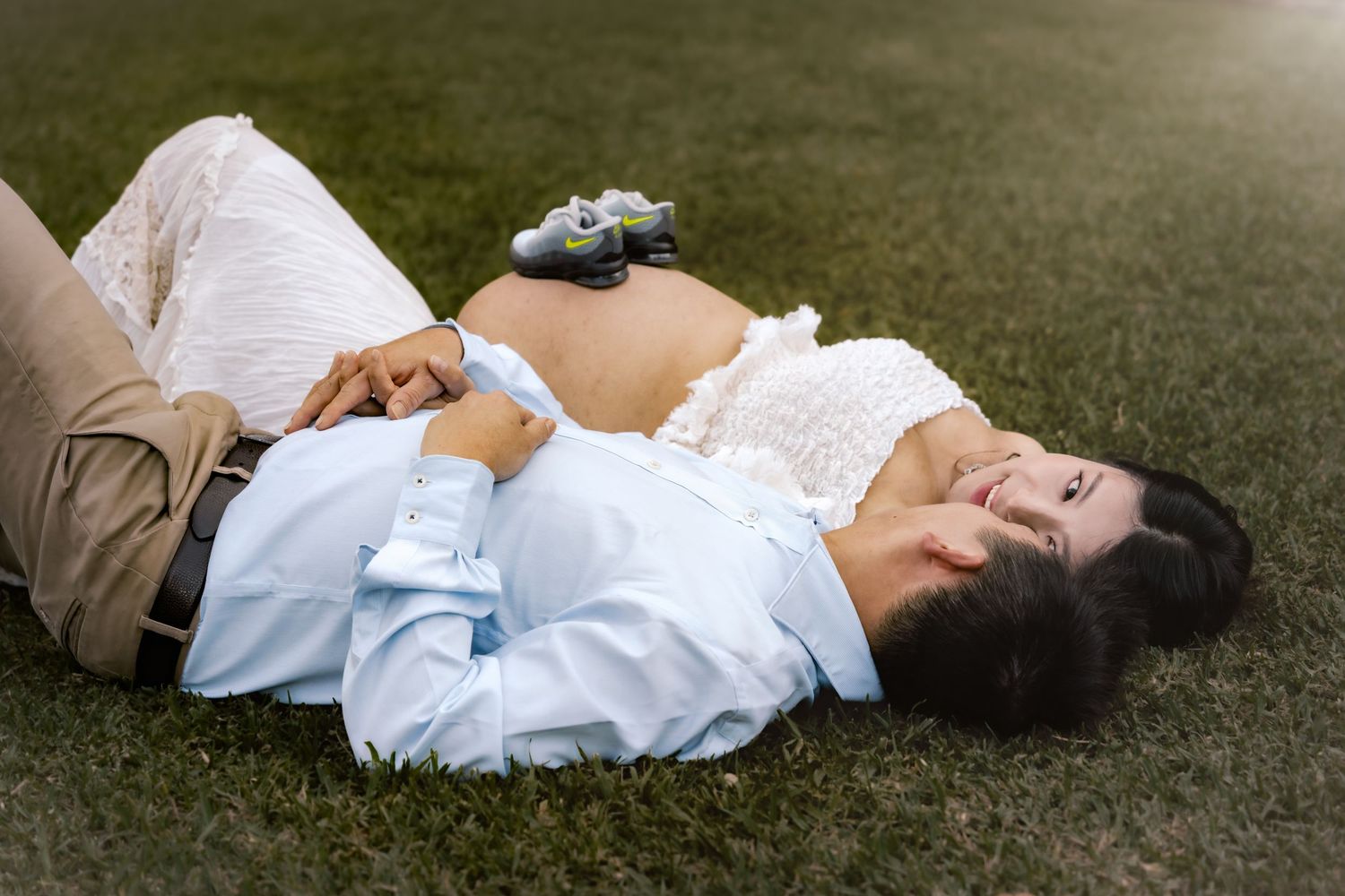 Pregnant couple lying on grass, woman resting on man's chest with baby shoes on her belly.