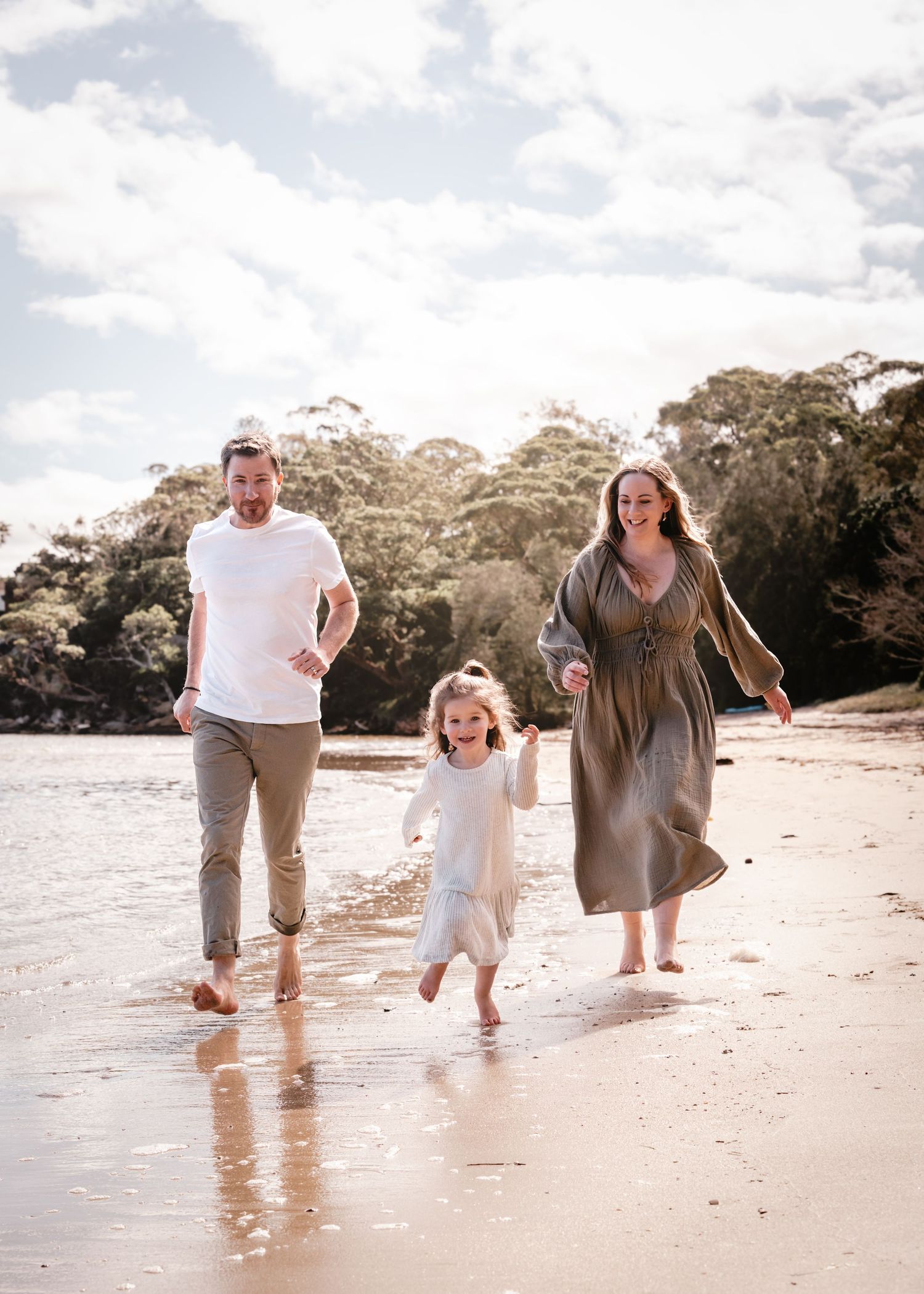 Three people playfully running and splashing through shallow ocean water at the beach on a sunny day.