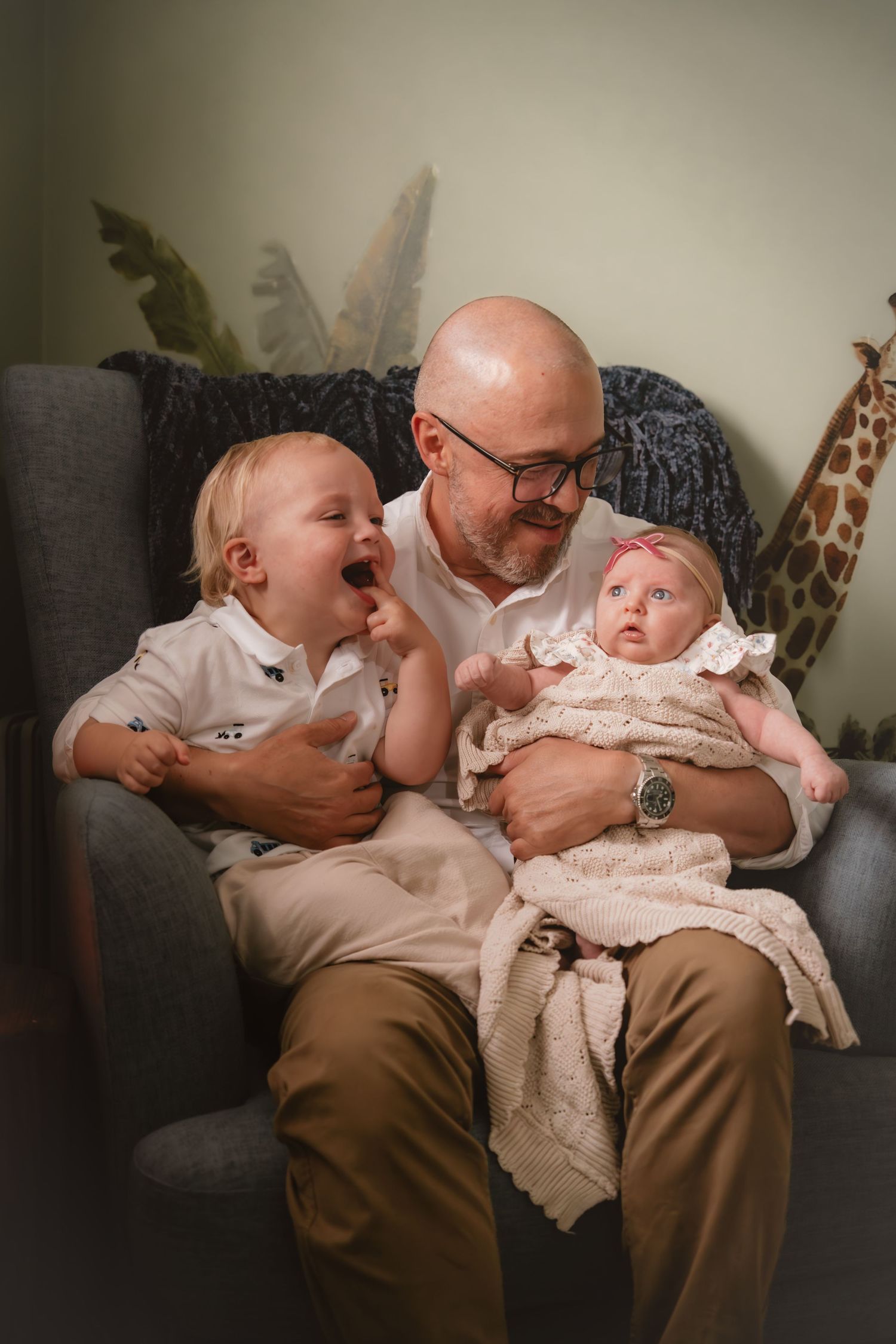 Bald father with glasses sits in gray chair holding newborn baby and laughing toddler in nursery room.