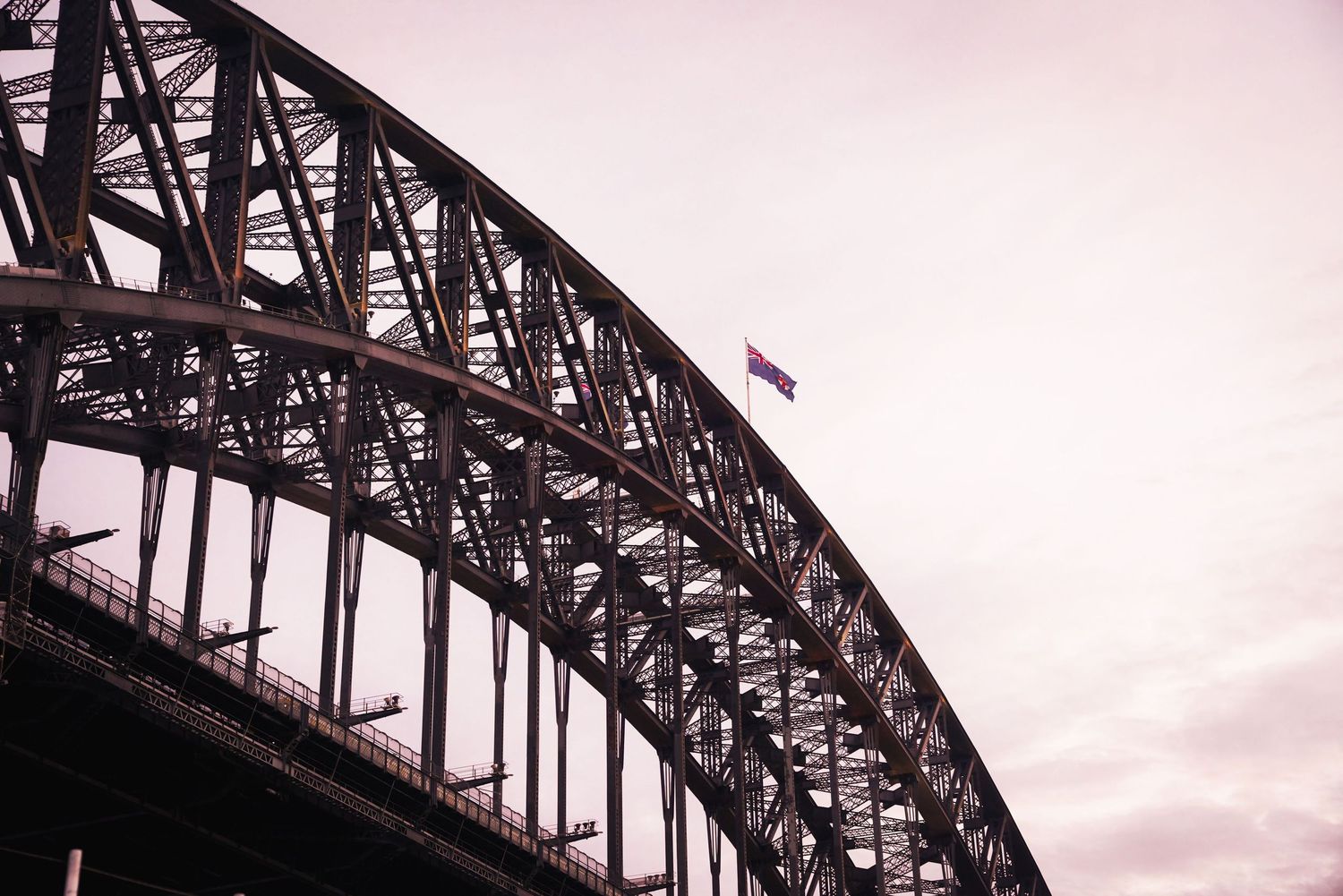 The iconic Sydney Harbour Bridge spans across the water at sunset with its distinctive steel arch silhouetted against the sky.