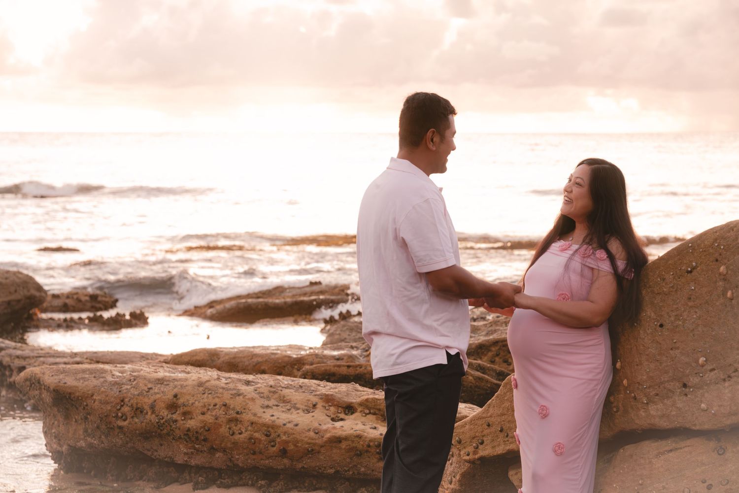 Couple holding hands on rocky beach at sunset, pregnant woman in pink dress smiling at partner in white shirt.