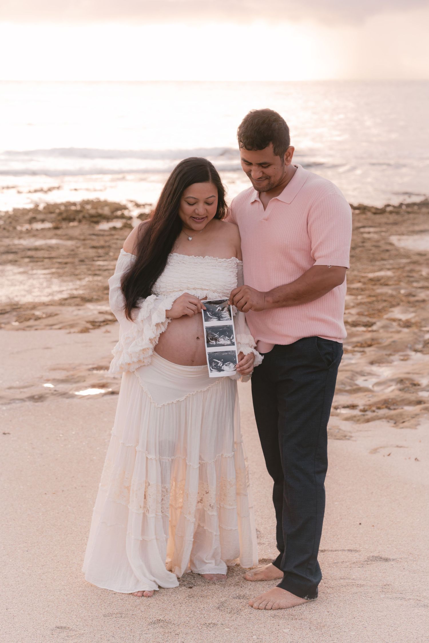 Expecting couple stands on beach at sunset, holding ultrasound photos while admiring pregnant belly in white dress.