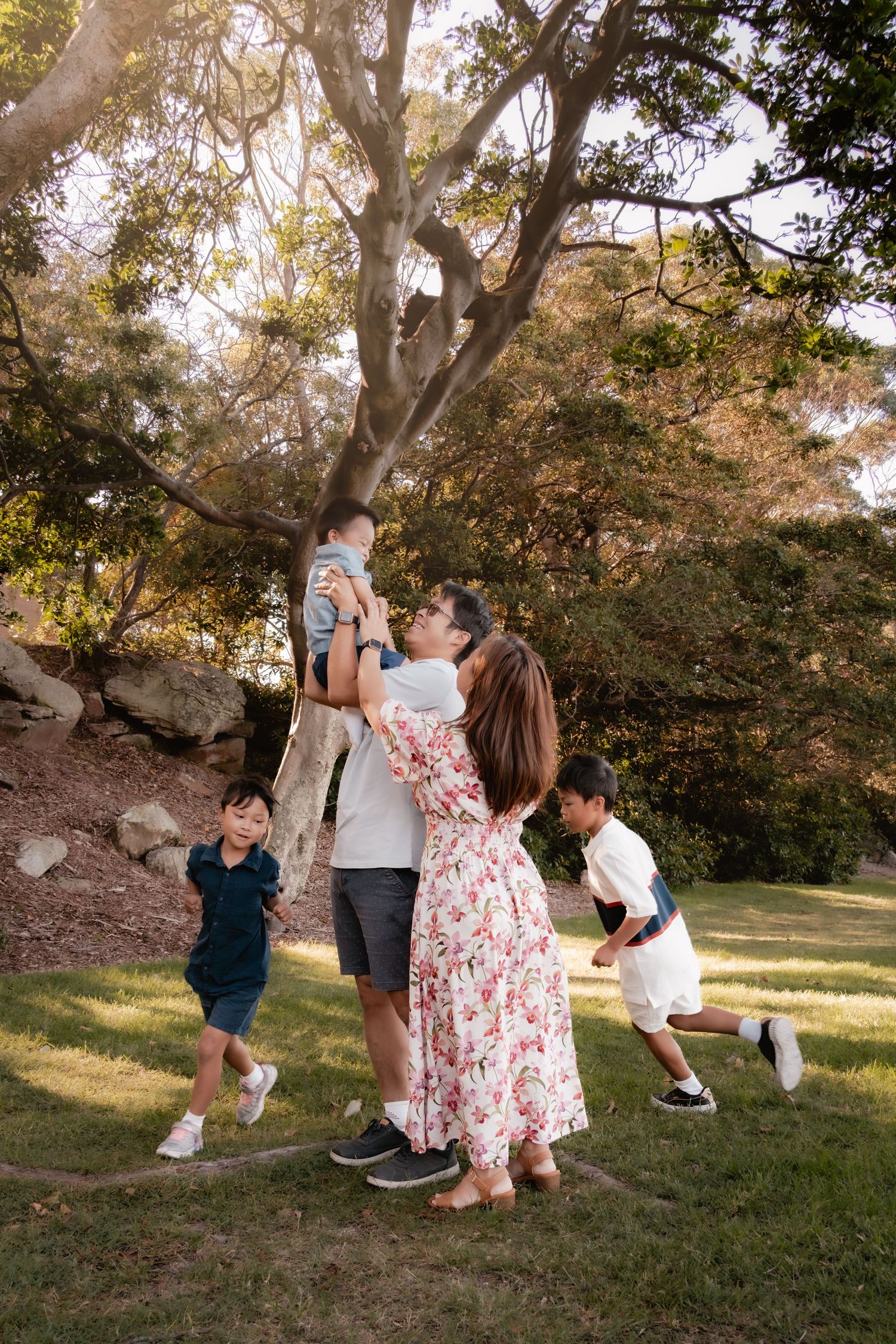 Family enjoying a sunny day in the park, with parents lifting a toddler while two children play around them.