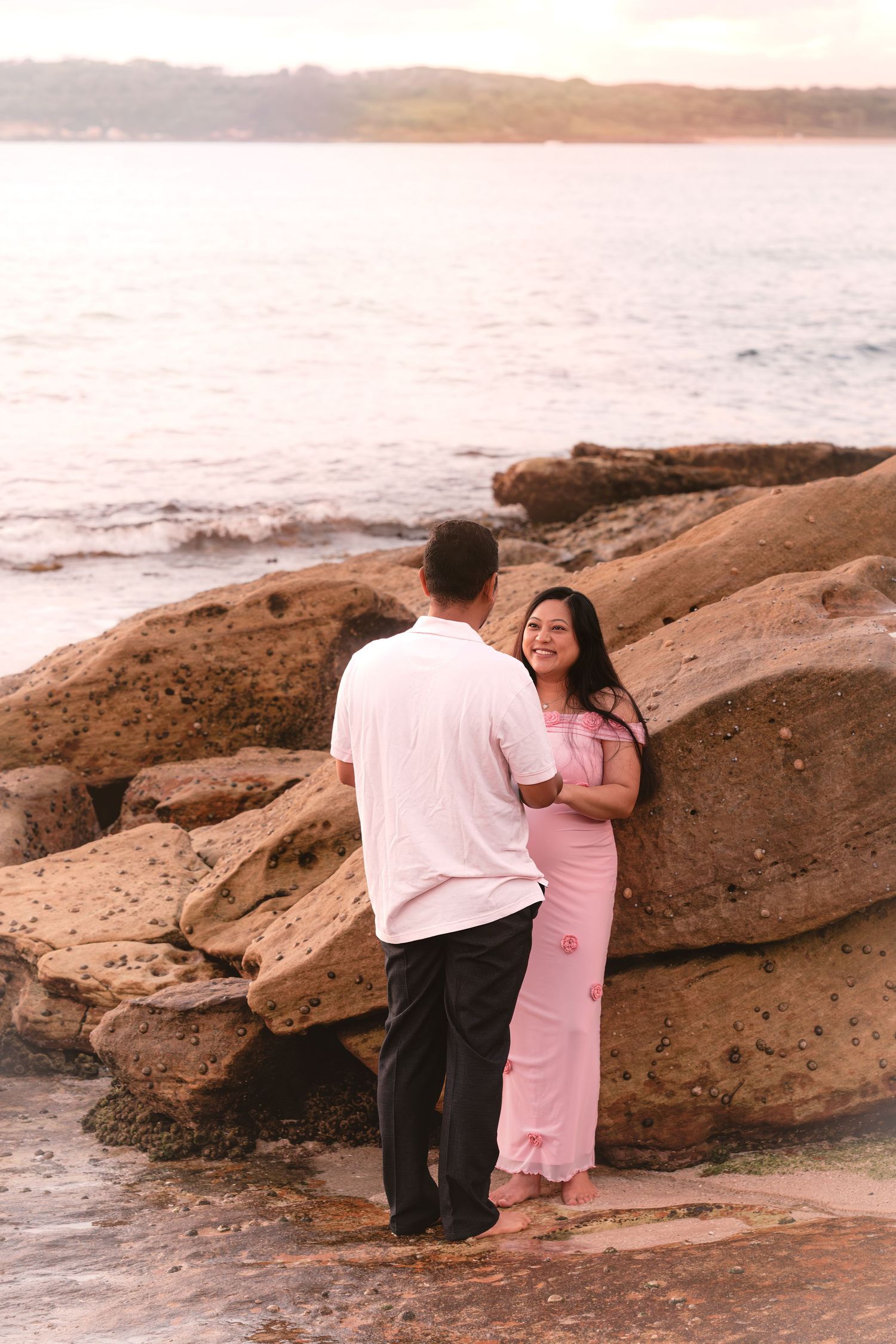 Couple in romantic pose on rocky beach at sunset, woman in pink dress smiling, man in white shirt and black pants.