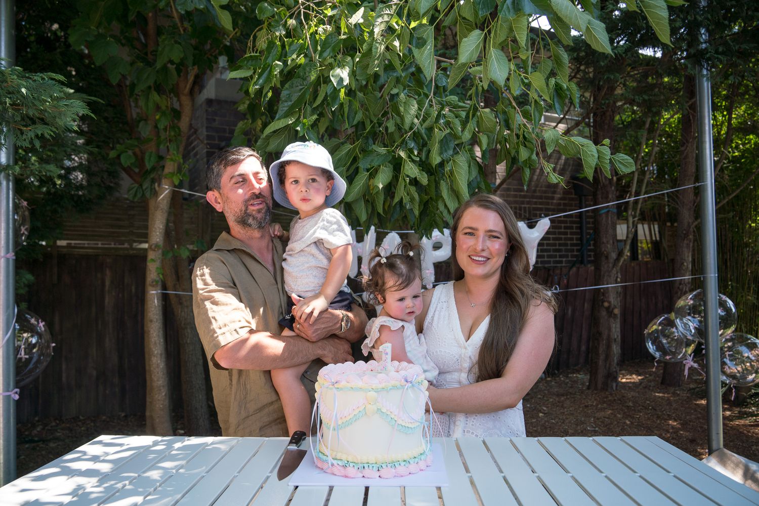 Family of four celebrating a first birthday outdoors with a white cake on a wooden table surrounded by lush greenery.