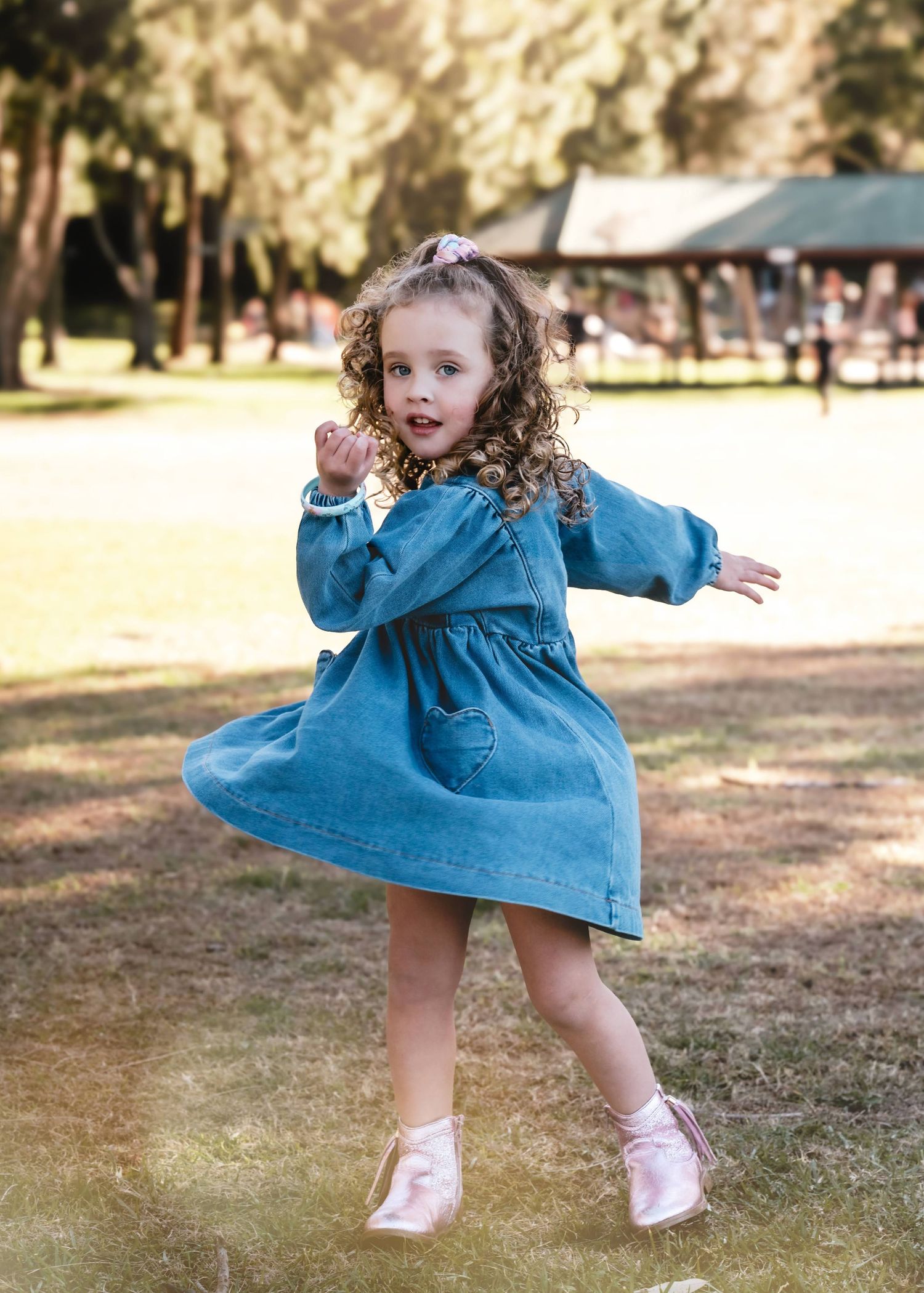 A small child in a blue denim dress twirls and dances playfully in a sunlit park.