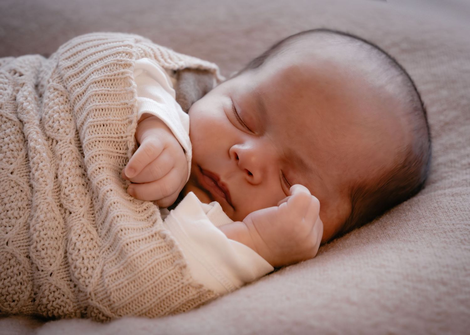 Sleeping baby in cozy beige knit sweater rests peacefully on soft gray bedding.