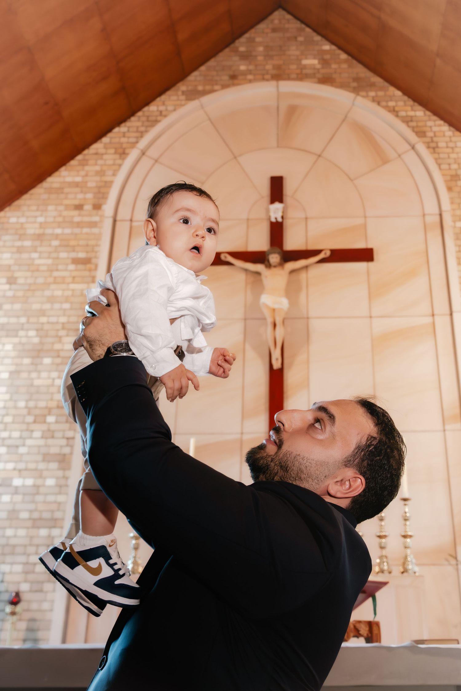 A bearded man in a black suit lifts a baby in white clothing inside a church with a crucifix in the background.