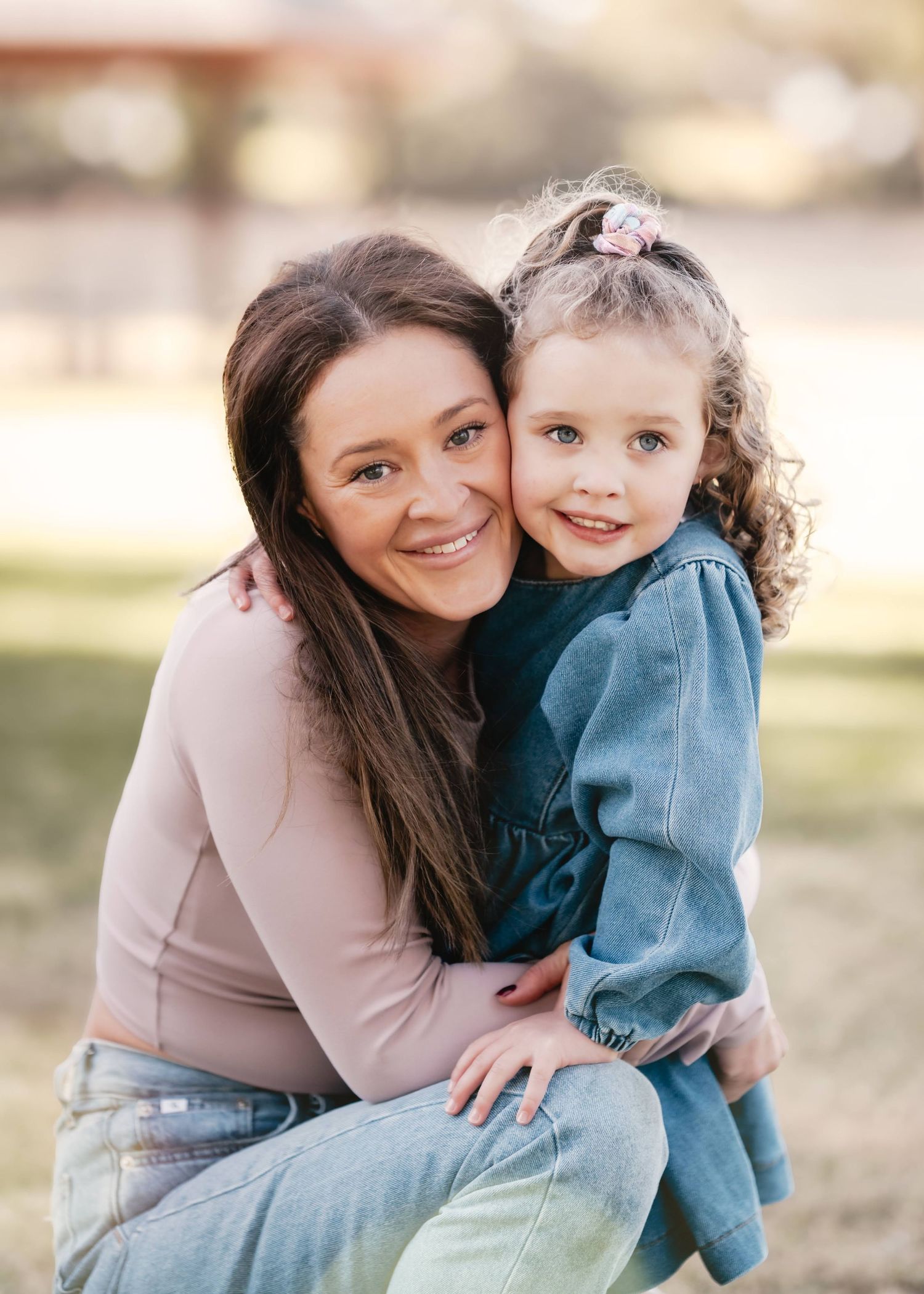 A warm outdoor family portrait shows a mother and daughter embracing while wearing casual clothing in soft natural light.