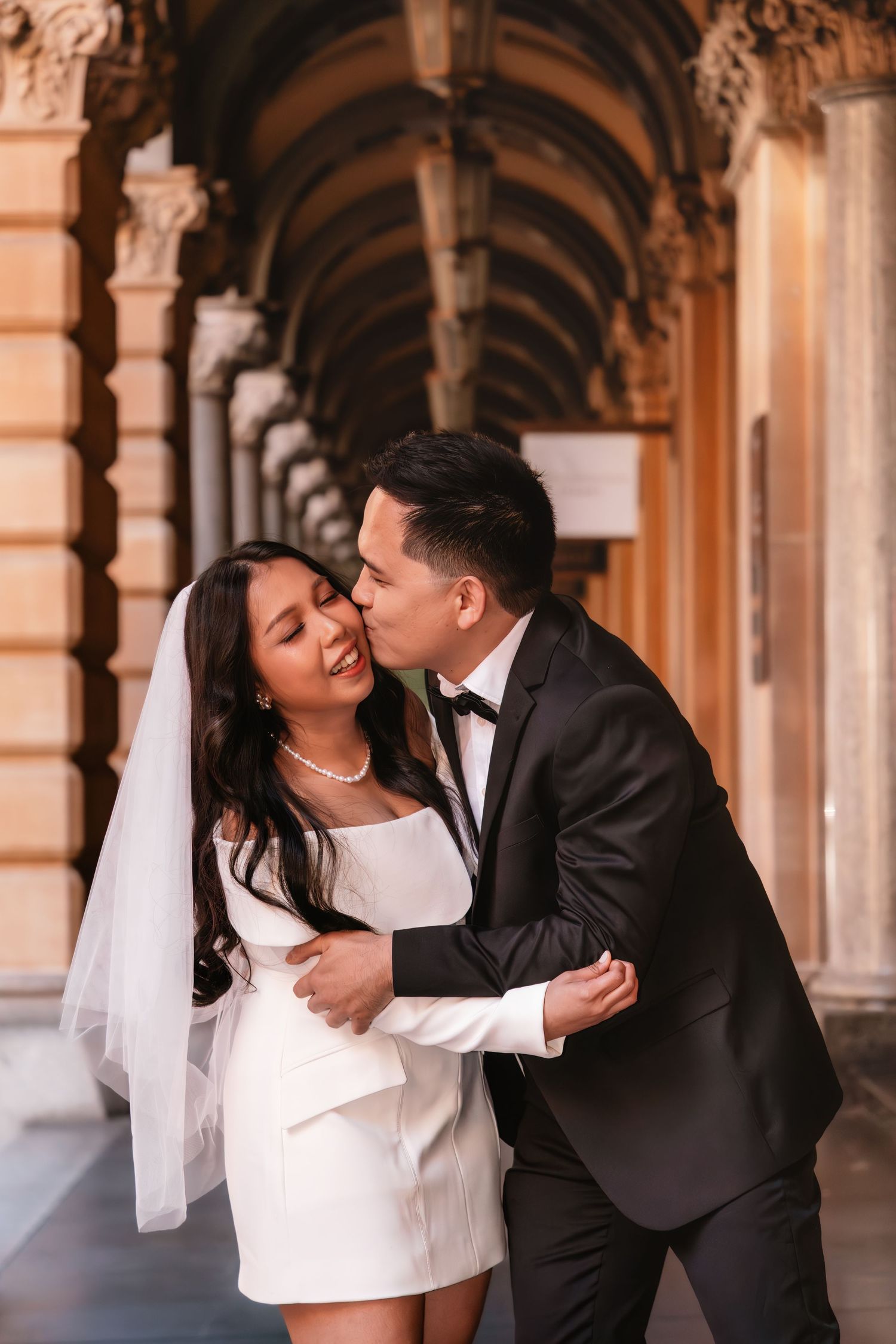 Bride and groom sharing a romantic kiss under arched colonnade, bride in white mini dress with veil.
