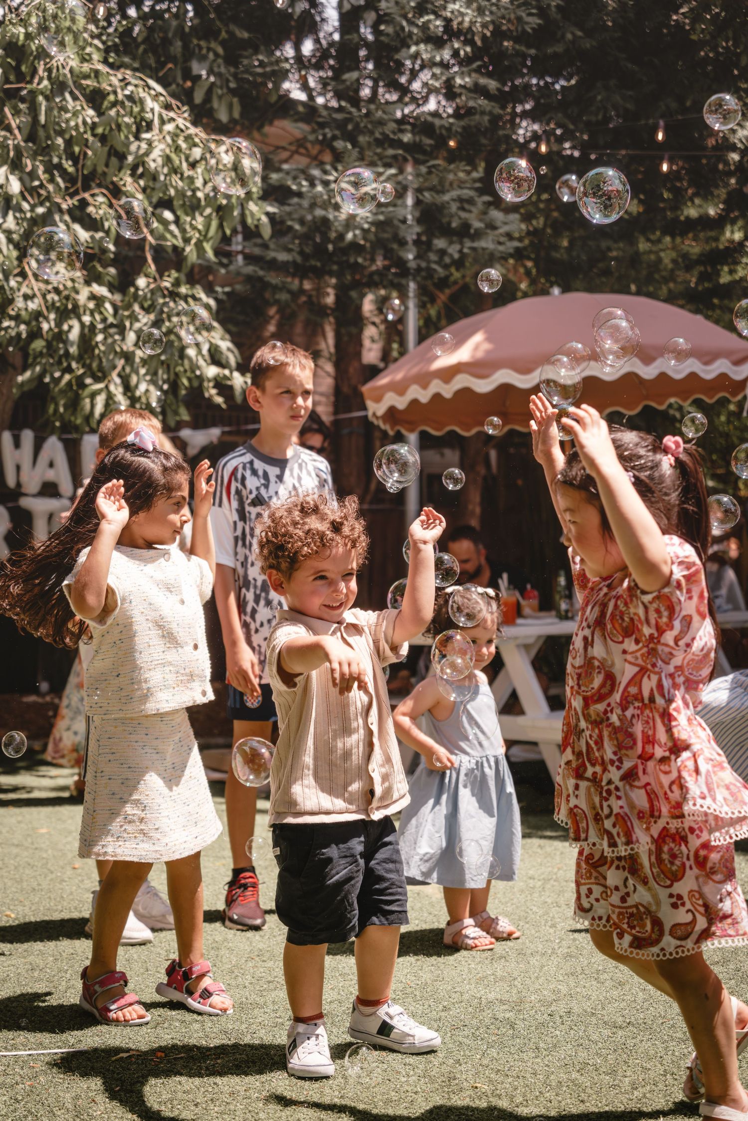 Children playing with bubbles outdoors at a birthday party, laughing and reaching up to catch floating soap bubbles.