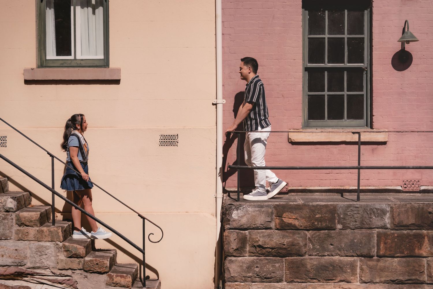 Two people stand on stone steps outside colorful buildings on a sunny day.